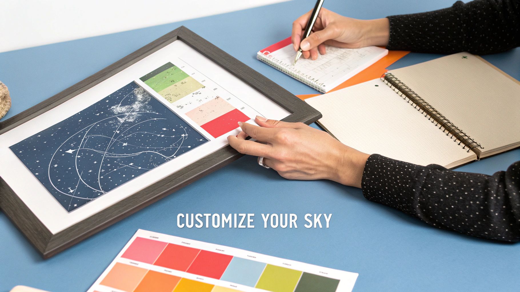 A person customizes a framed star map print at a desk with notebooks and color swatches.