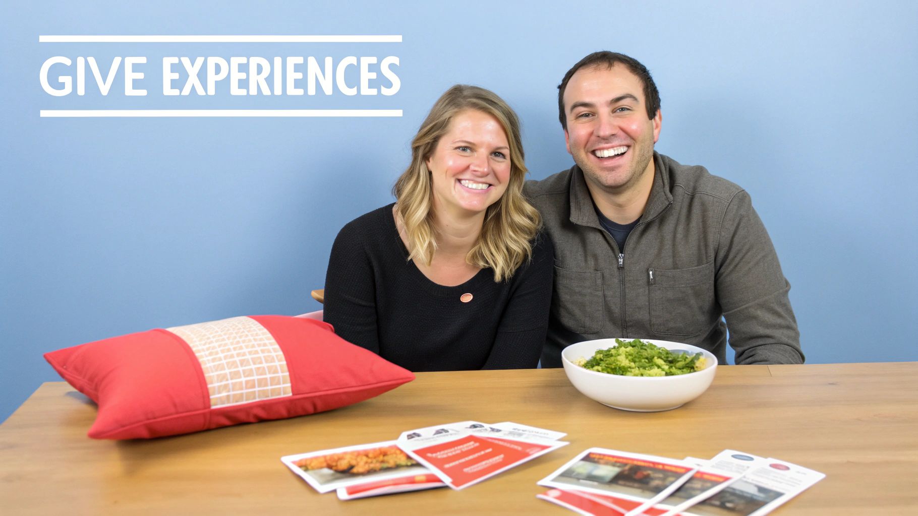 A smiling man and woman behind a table with a bowl of food, gift cards, and "GIVE EXPERIENCES" on the wall.