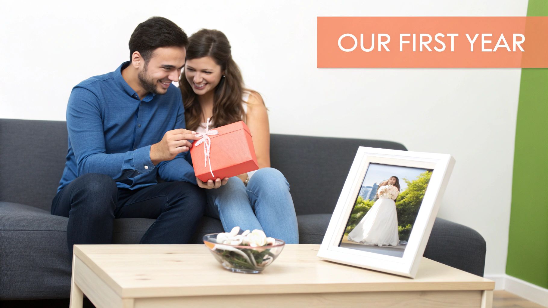 A happy couple on a sofa, the man opening a red gift box, with a framed wedding photo on a table.