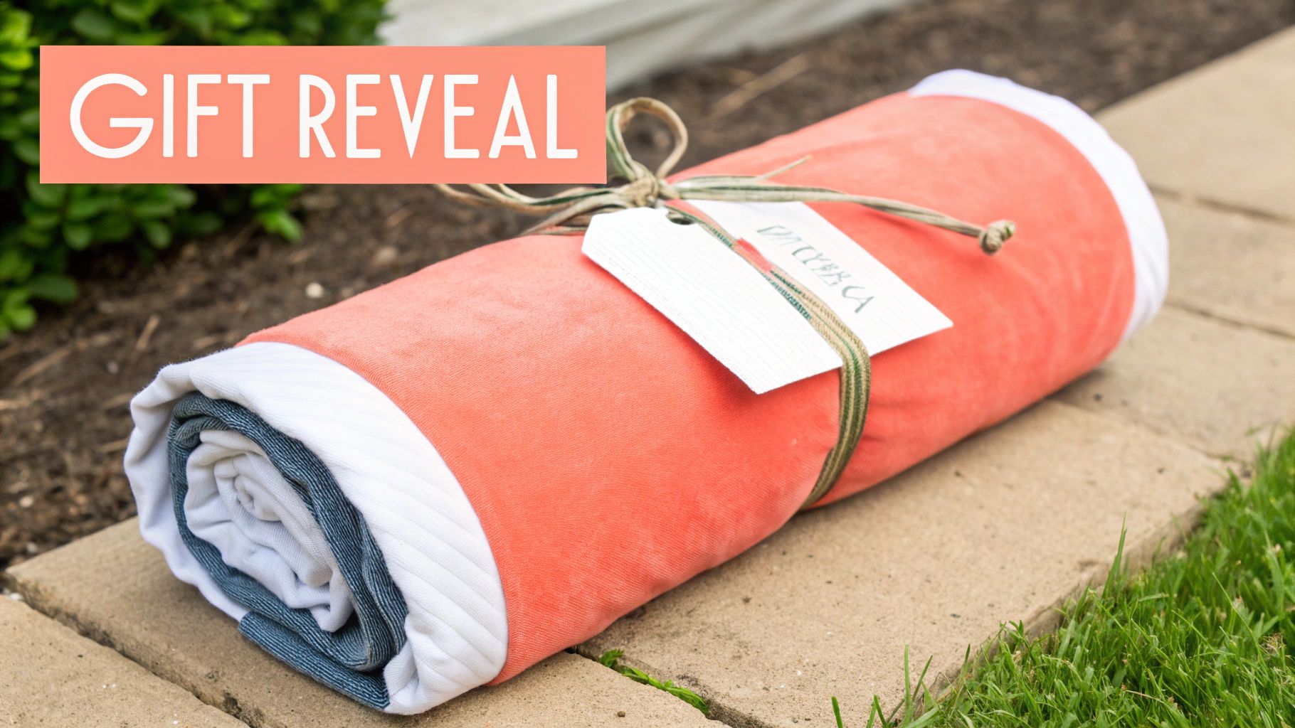 A neatly rolled coral blanket with a personalized gift tag sits on concrete pavers outdoors.