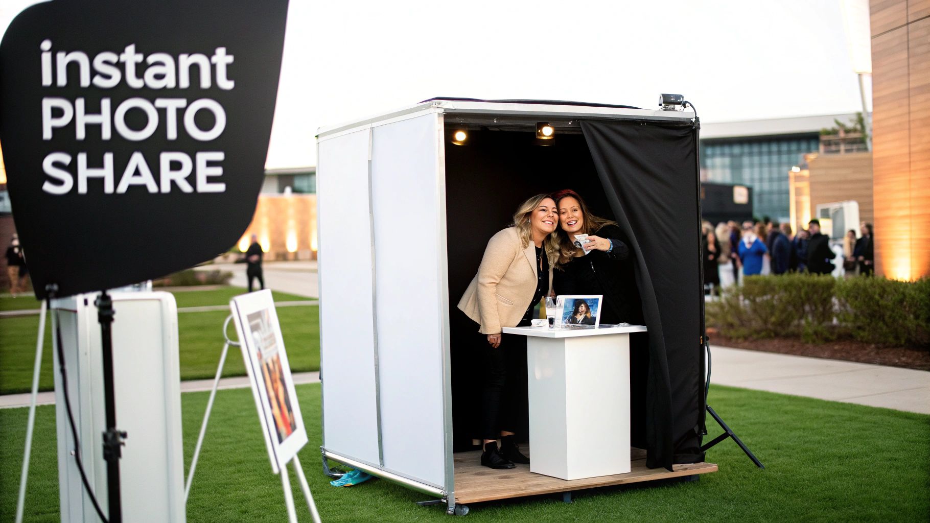 Two happy women smiling while taking a selfie in a portable outdoor instant photo booth at an event.