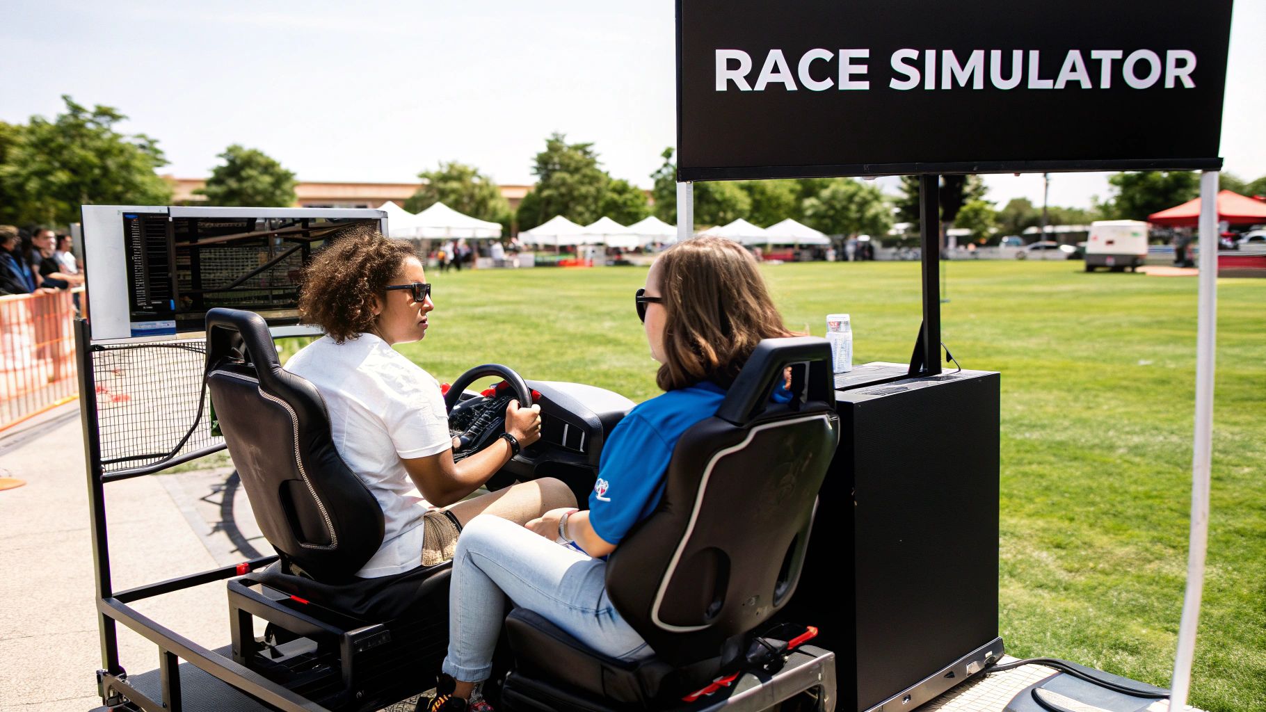 Two women wearing sunglasses play a race simulator game outdoors on a sunny day.