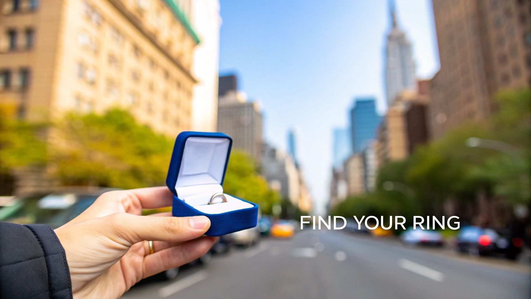 A hand holds an open blue velvet ring box with a silver engagement ring on a New York City street.