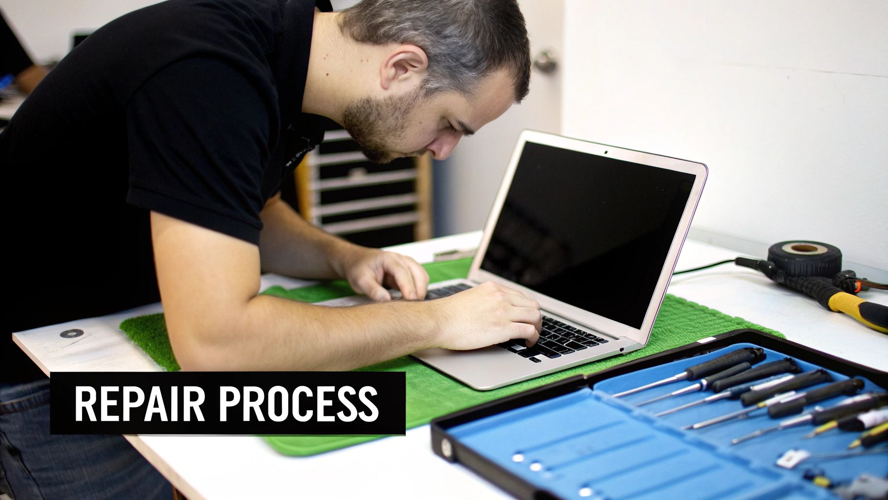 A bearded man working on a silver MacBook Air laptop, with a blue toolkit open on the table.