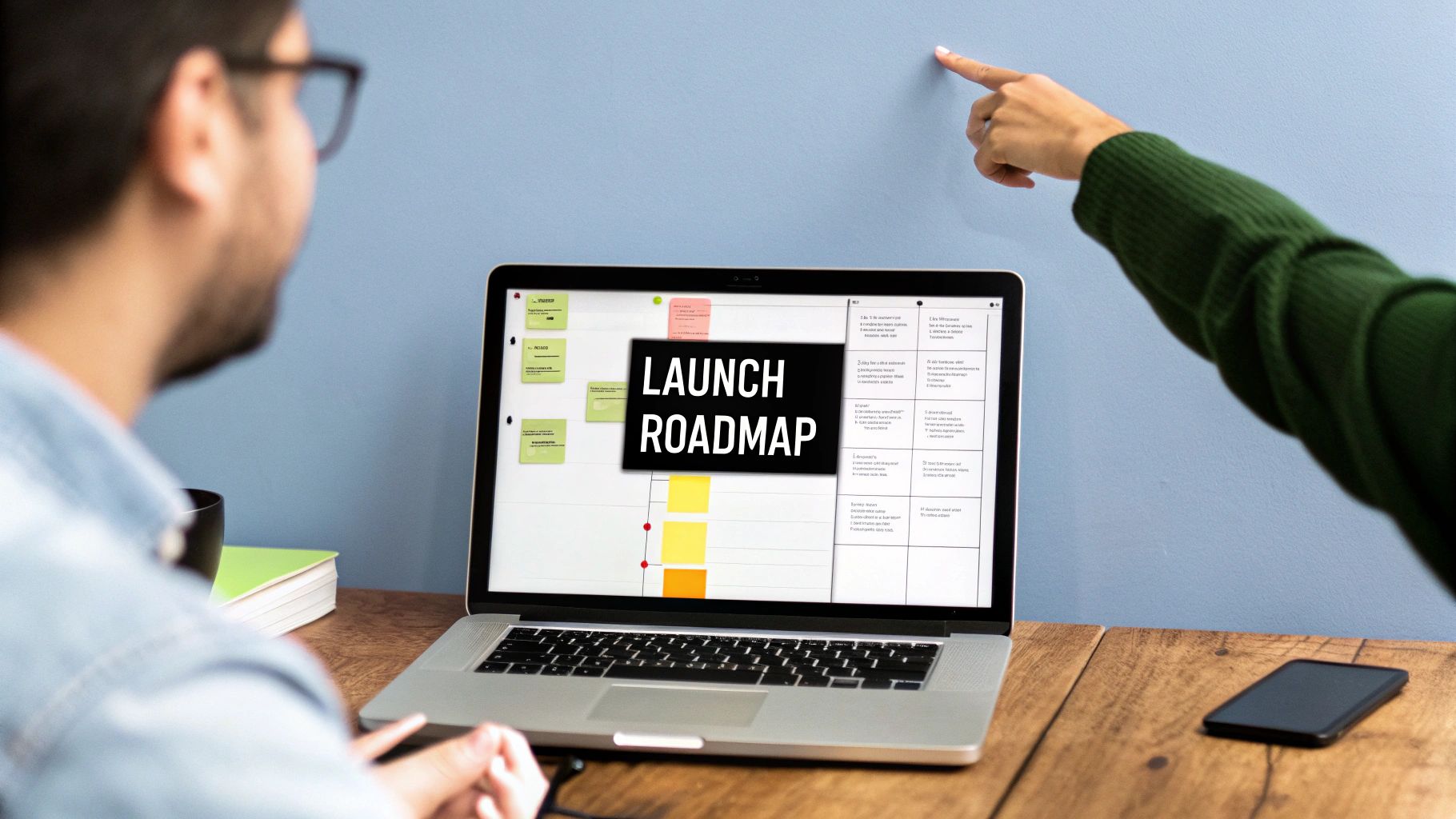 Two people collaborating on a launch roadmap displayed on a laptop screen on a wooden desk.