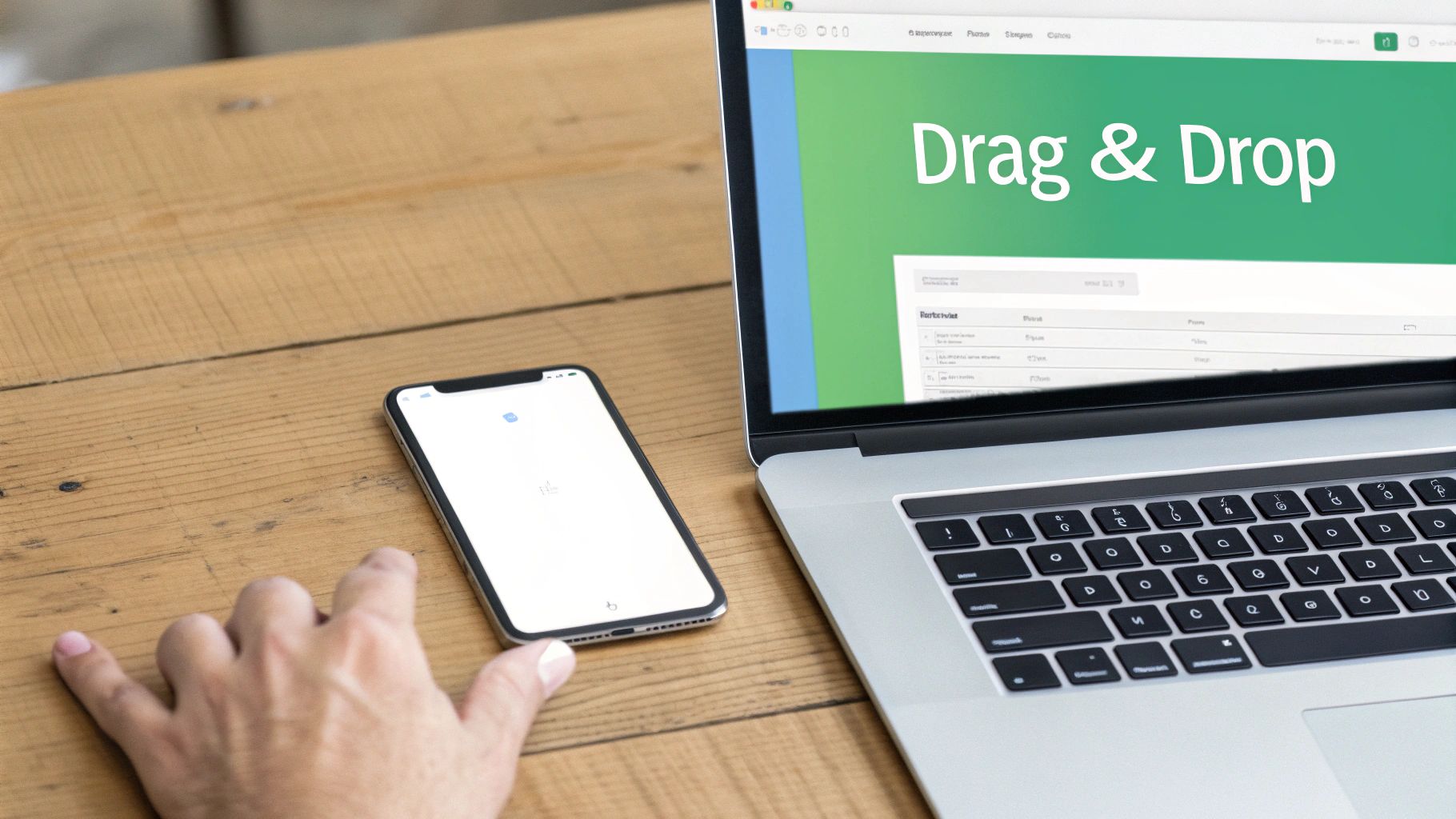 A person's hand next to a smartphone and laptop on a wooden desk, with "Drag & Drop" on the laptop screen.