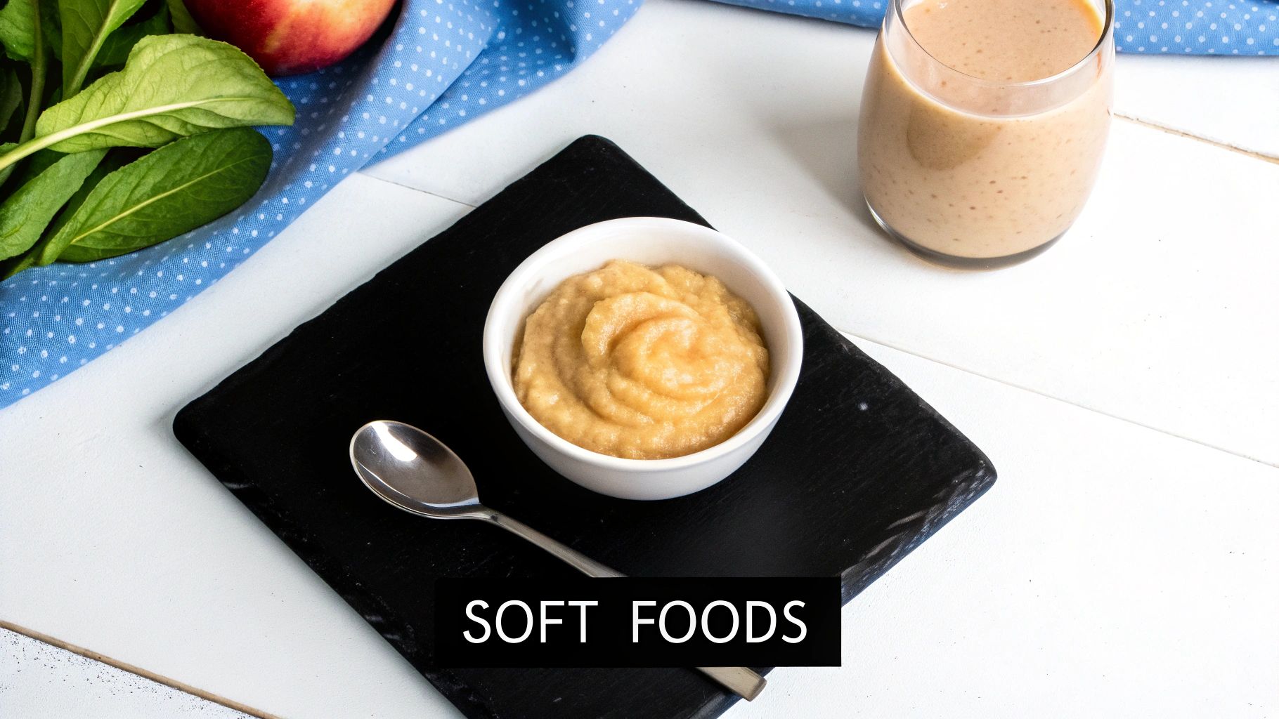 Overhead shot of a white bowl of applesauce, a spoon, a smoothie, and fruit on a table, perfect for soft foods.