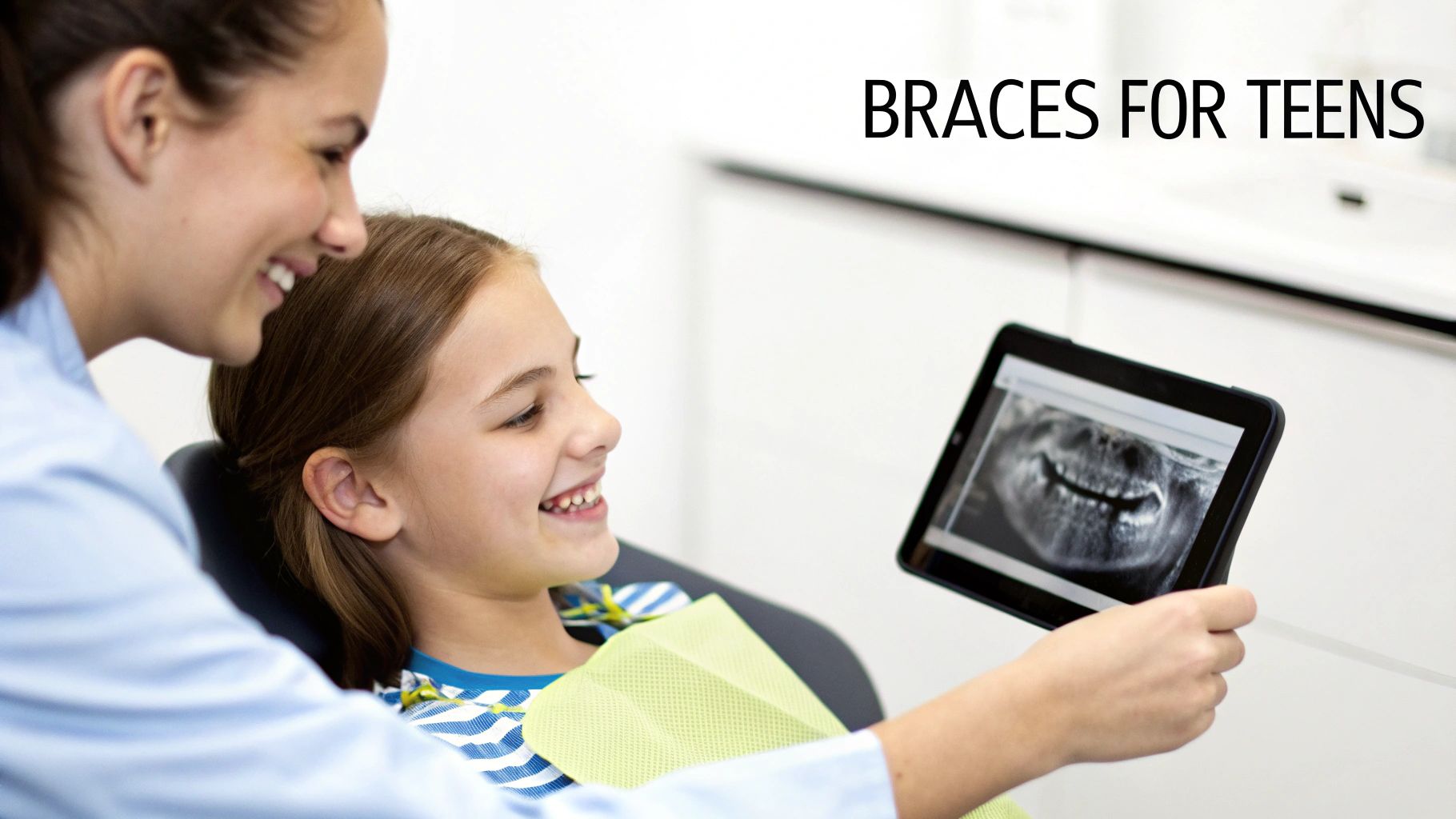 A smiling dentist shows a young girl her dental X-ray on a tablet, discussing braces.