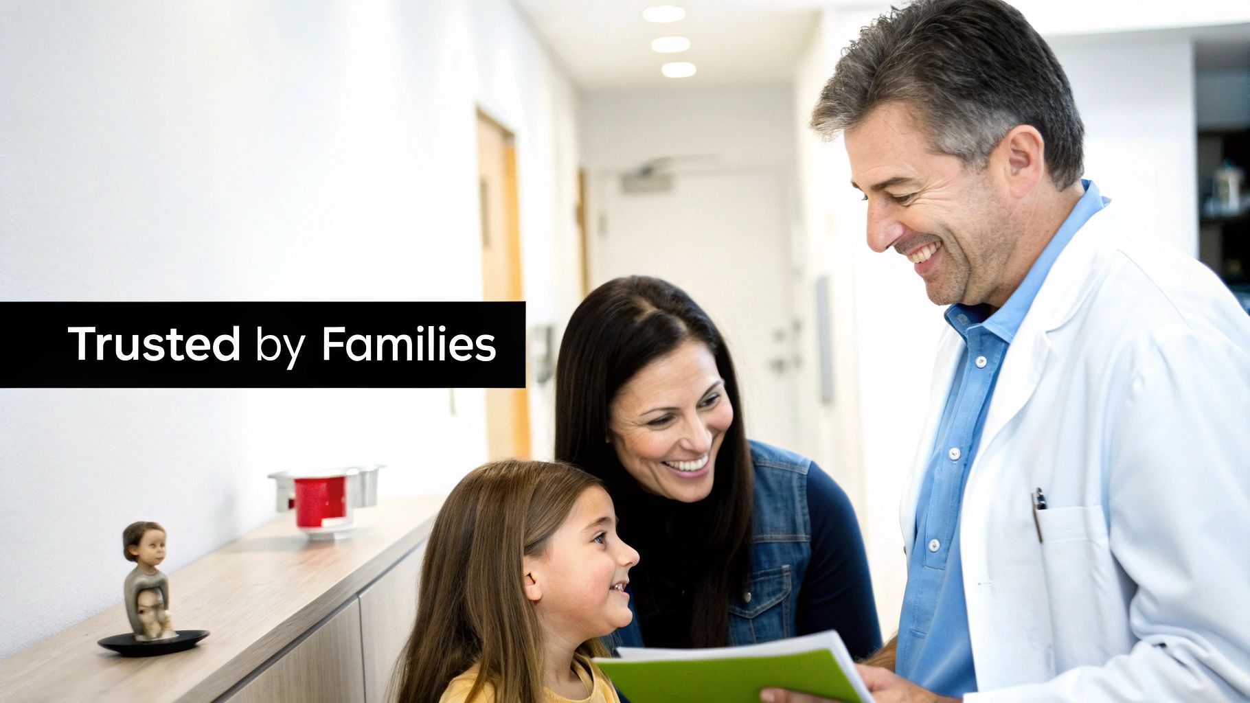 A smiling male doctor in a white coat talks to a mother and her young daughter holding a green folder.