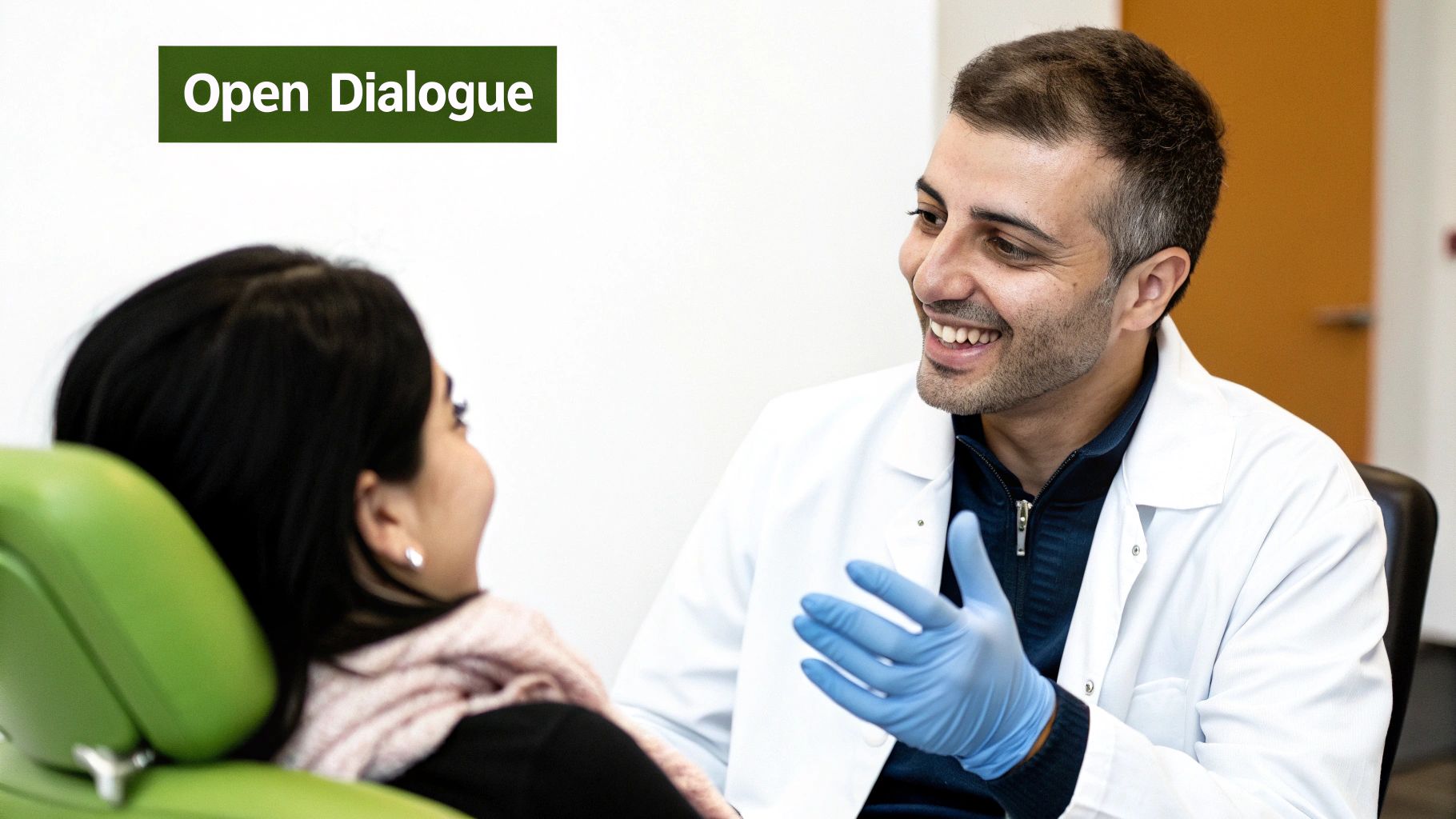 Smiling dentist in a white coat and blue gloves engaging in open dialogue with a patient in a green dental chair.