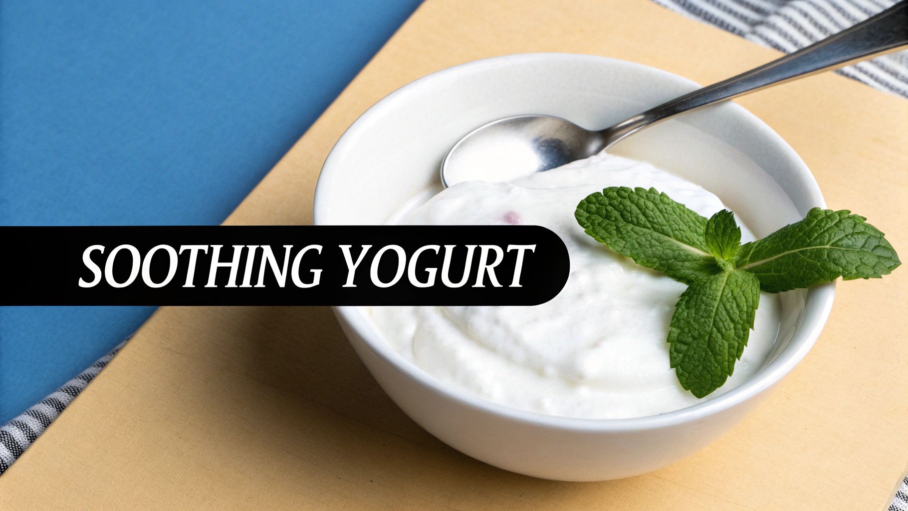 A white bowl of creamy yogurt topped with fresh mint leaves and a spoon, on a wooden and blue surface.