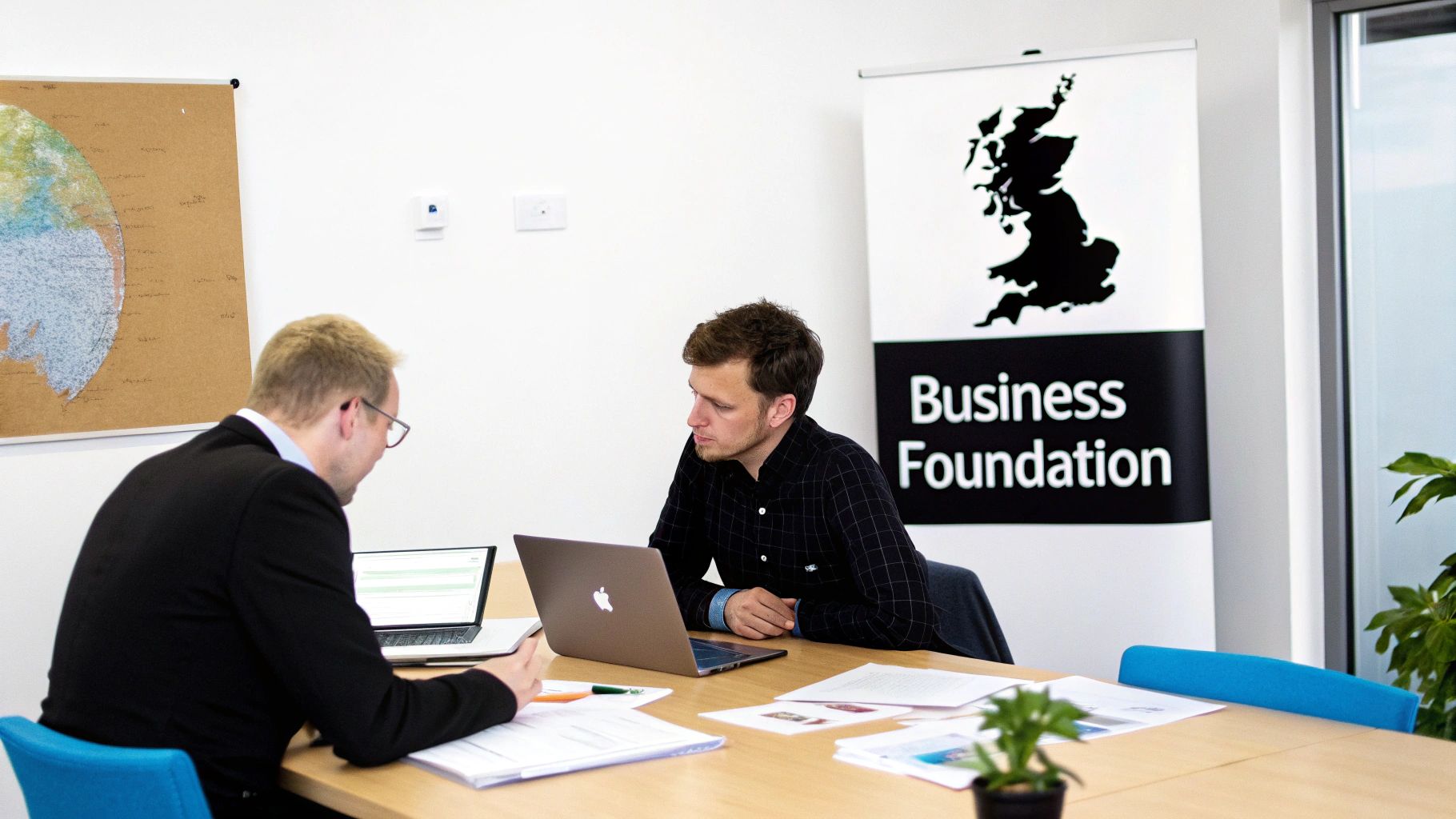 Two men in an office, engaged in a business meeting with laptops and documents on a wooden table.