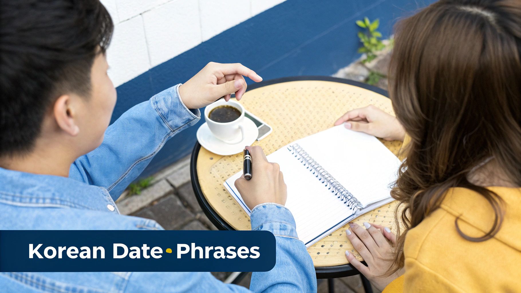 Two individuals studying Korean date phrases at an outdoor cafe, with coffee, a phone, and a notebook.