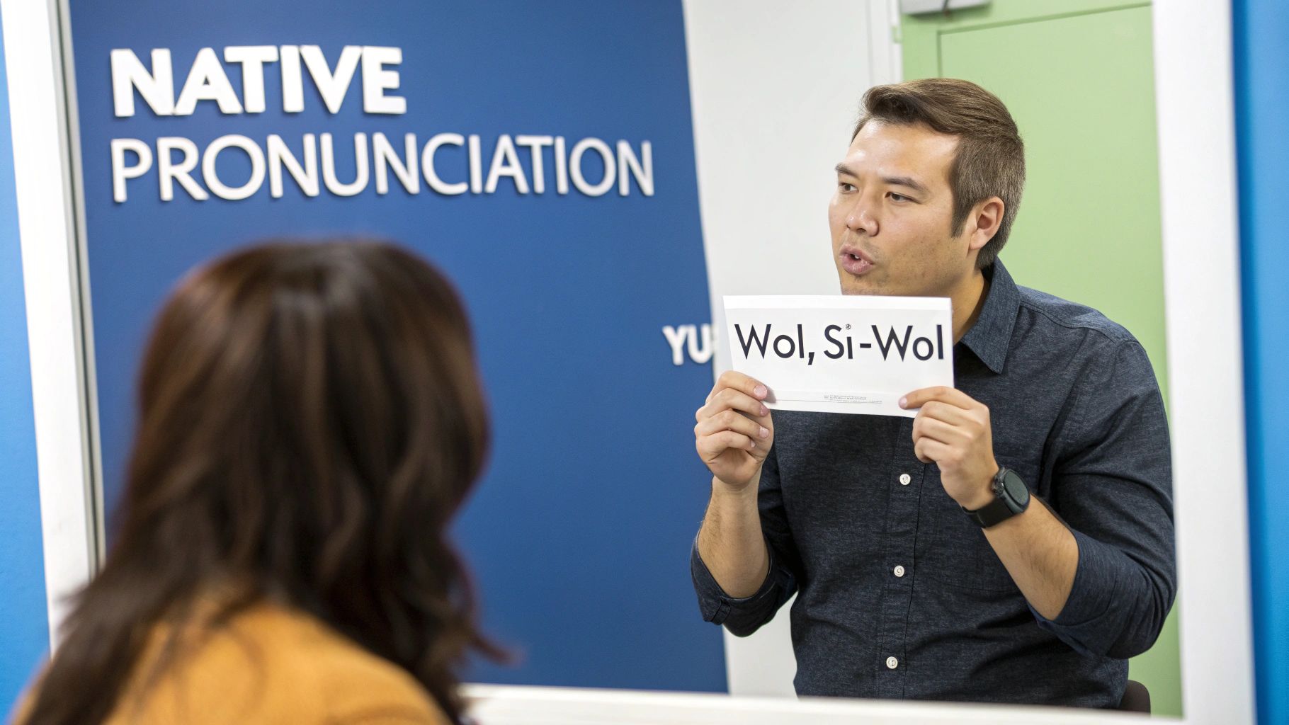 A man teaches Korean pronunciation, holding a sign with 'Wol, Si-Wol', to a student.