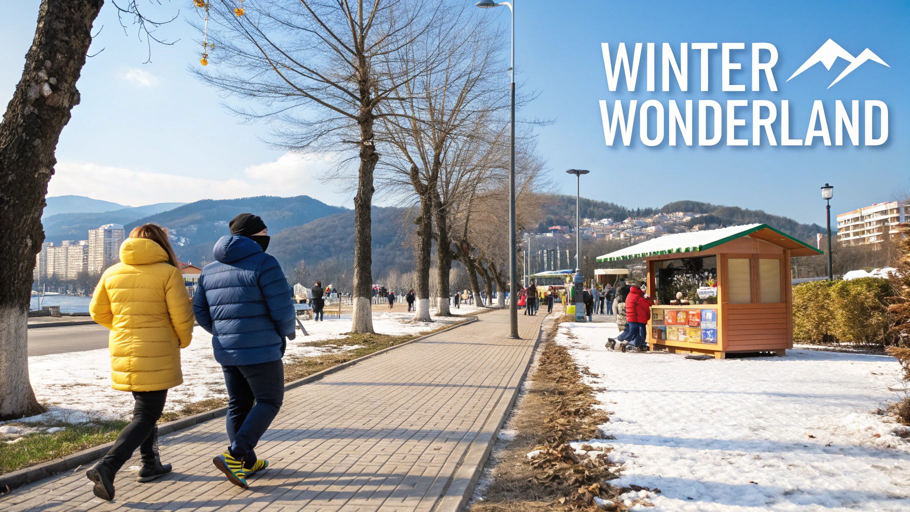Two people in winter coats walk on a snowy path by a lake with mountains and market stalls.