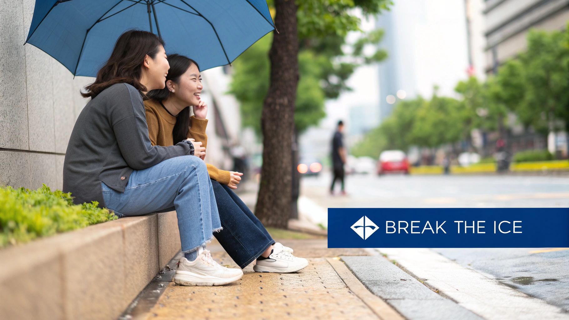 Two happy Asian women sit under a blue umbrella, laughing and talking on a rainy city street.