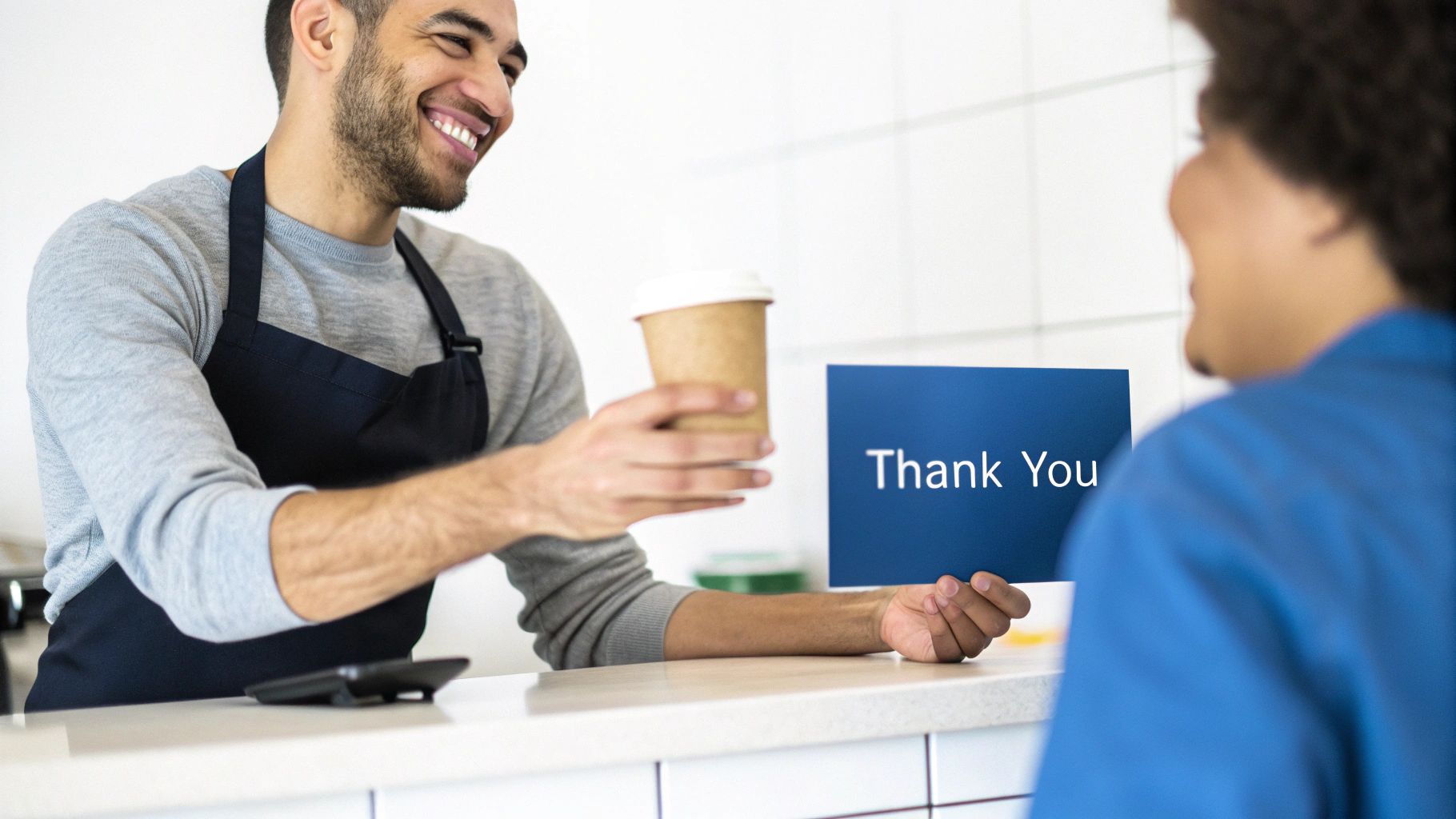 A happy barista in an apron hands a coffee cup to a customer, with a 'Thank You' sign visible.