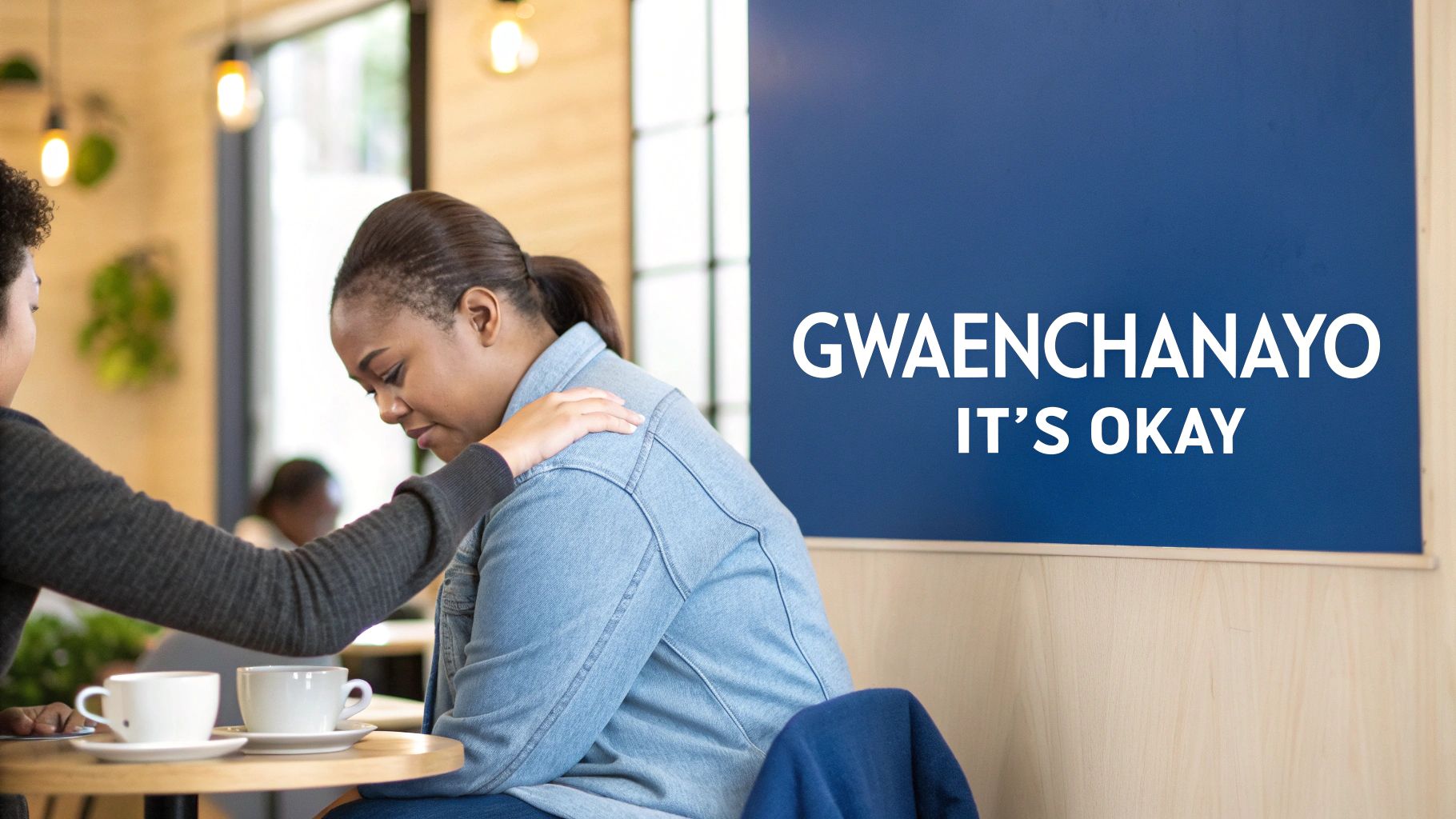 A woman comforts another sad woman in a cafe, with a blue sign reading 'IT'S OKAY' in the background.