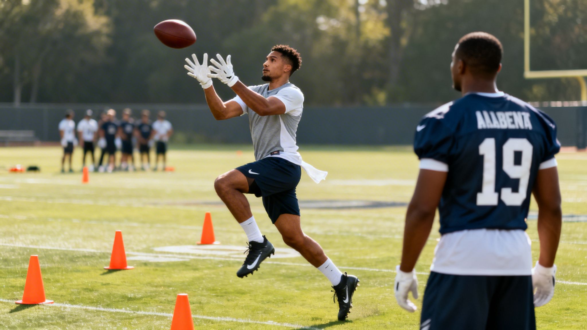 A football player in a grey shirt catches a ball on a sunny field during practice.