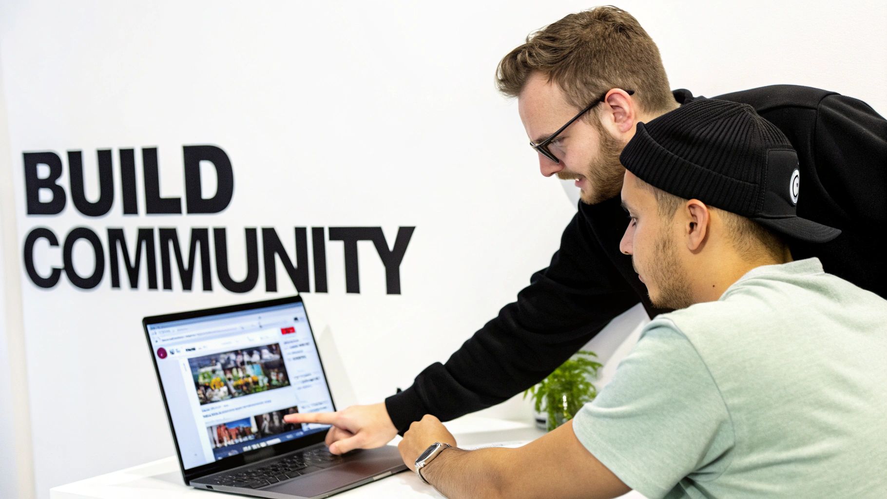 Two men collaborate on a laptop, viewing a social media feed with 'BUILD COMMUNITY' on the wall.