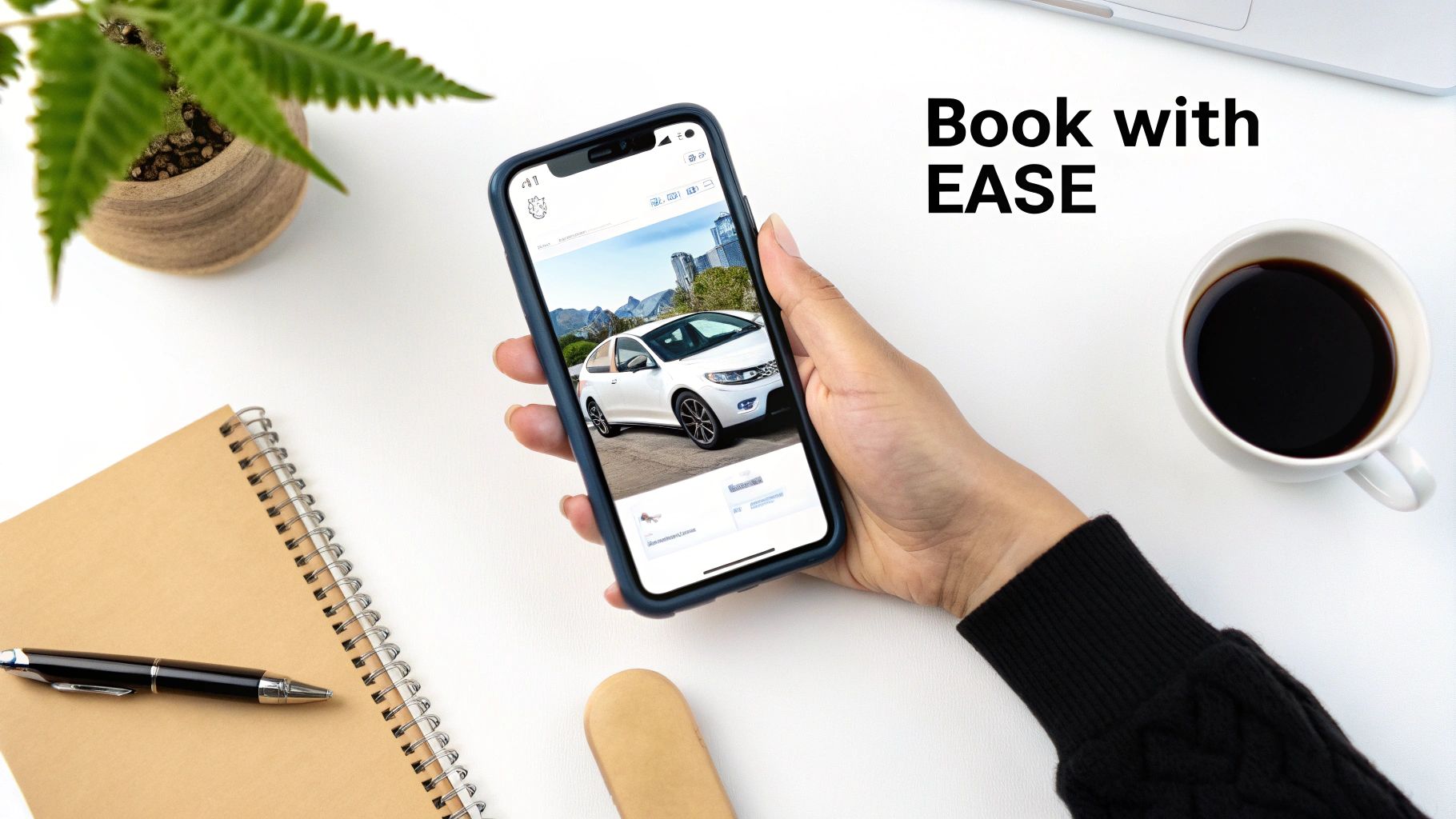 A person holds a smartphone showing a white car on screen, next to a coffee cup and notebook.