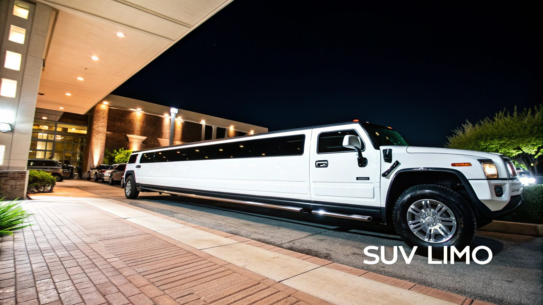 A sleek white SUV limousine parked at night outside a well-lit building entrance, ready for transport.