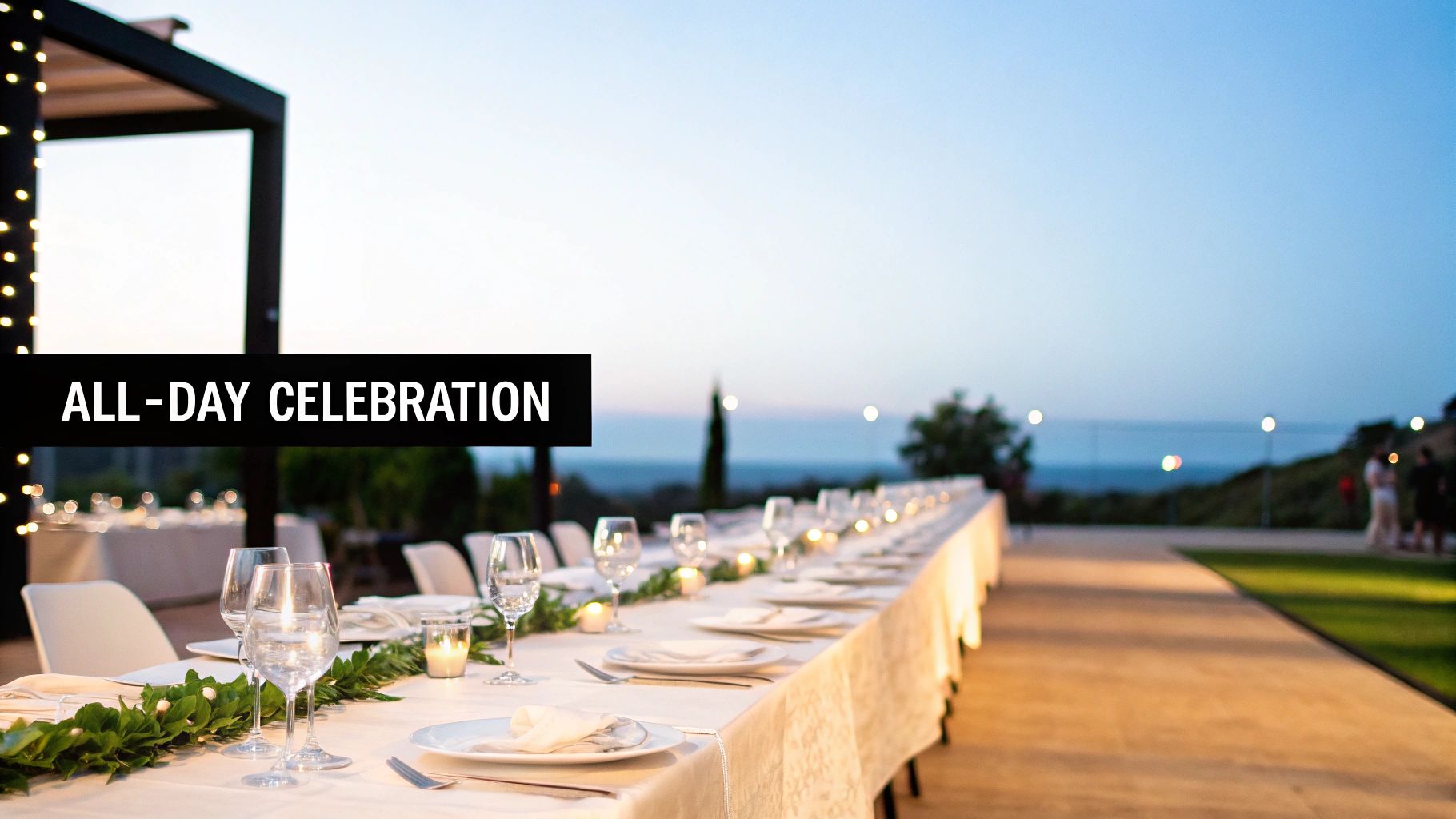 Elegant long outdoor dining table set for an evening celebration with fairy lights and a sea view.