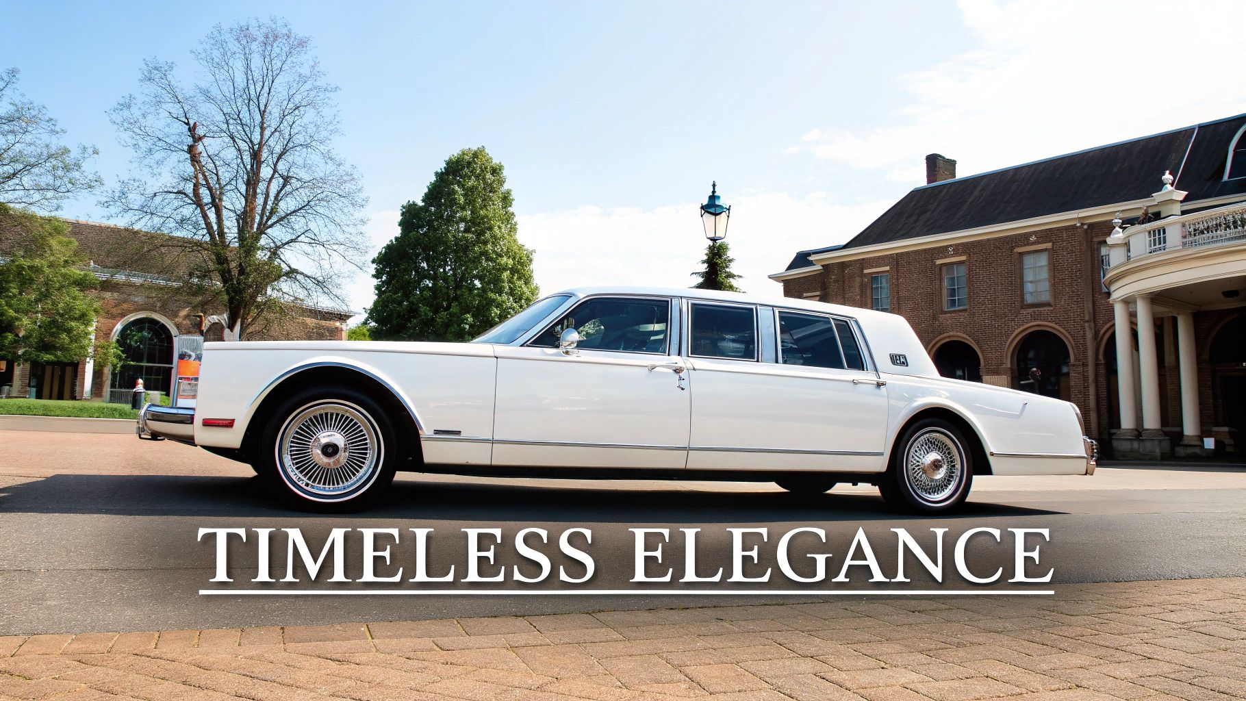 Elegant white vintage limousine parked by a classic brick building and trees on a sunny day.