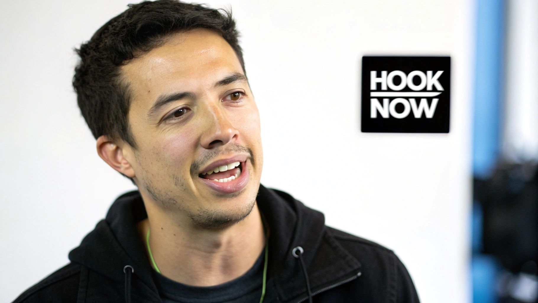 Close-up of a man with dark hair and stubble, wearing a black hoodie, talking happily. A 'HOOK NOW' sign is in the background.