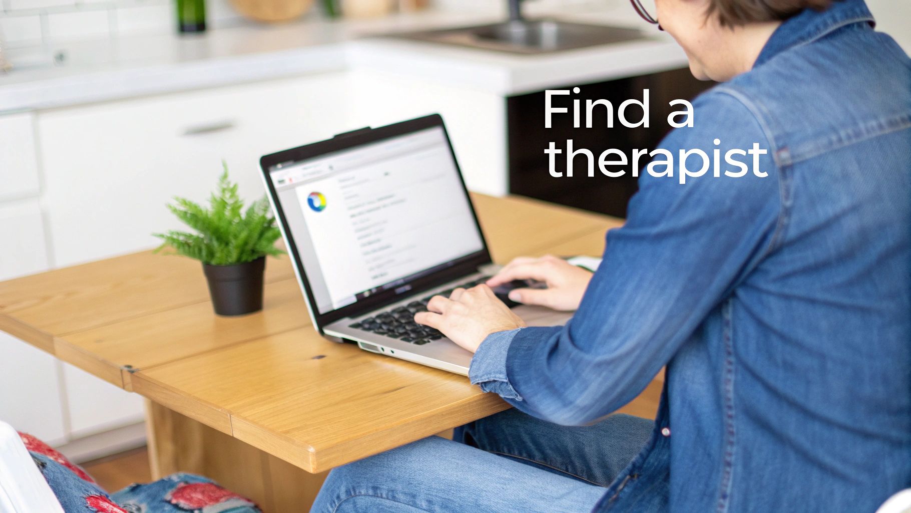 Person sitting at a wooden table, typing on a laptop to find a therapist online, with a plant.