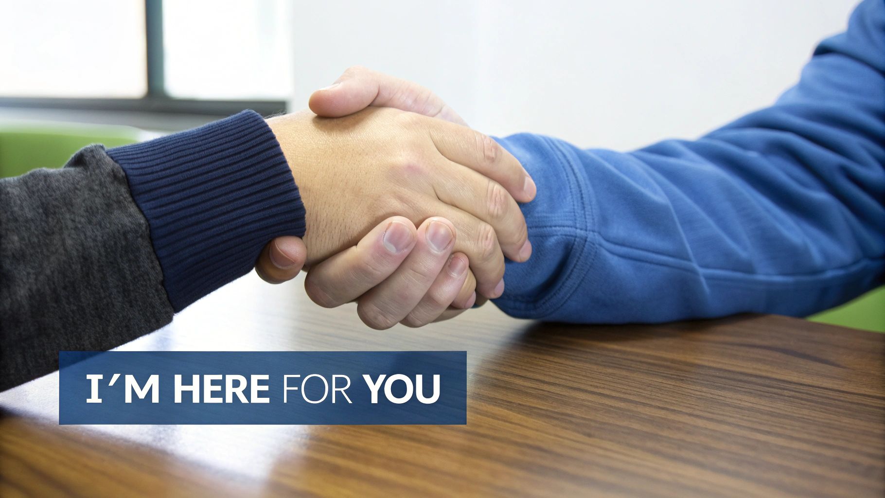 Two individuals in long-sleeved shirts shaking hands across a wooden desk, symbolizing support.