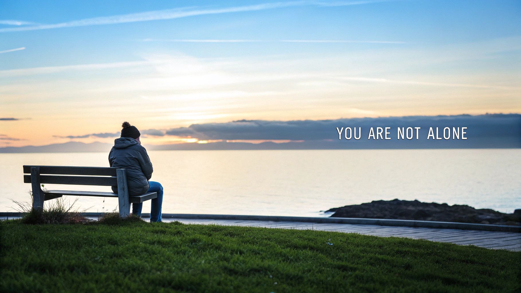 A person sits alone on a bench overlooking the ocean at sunset, with text 'YOU ARE NOT ALONE'.