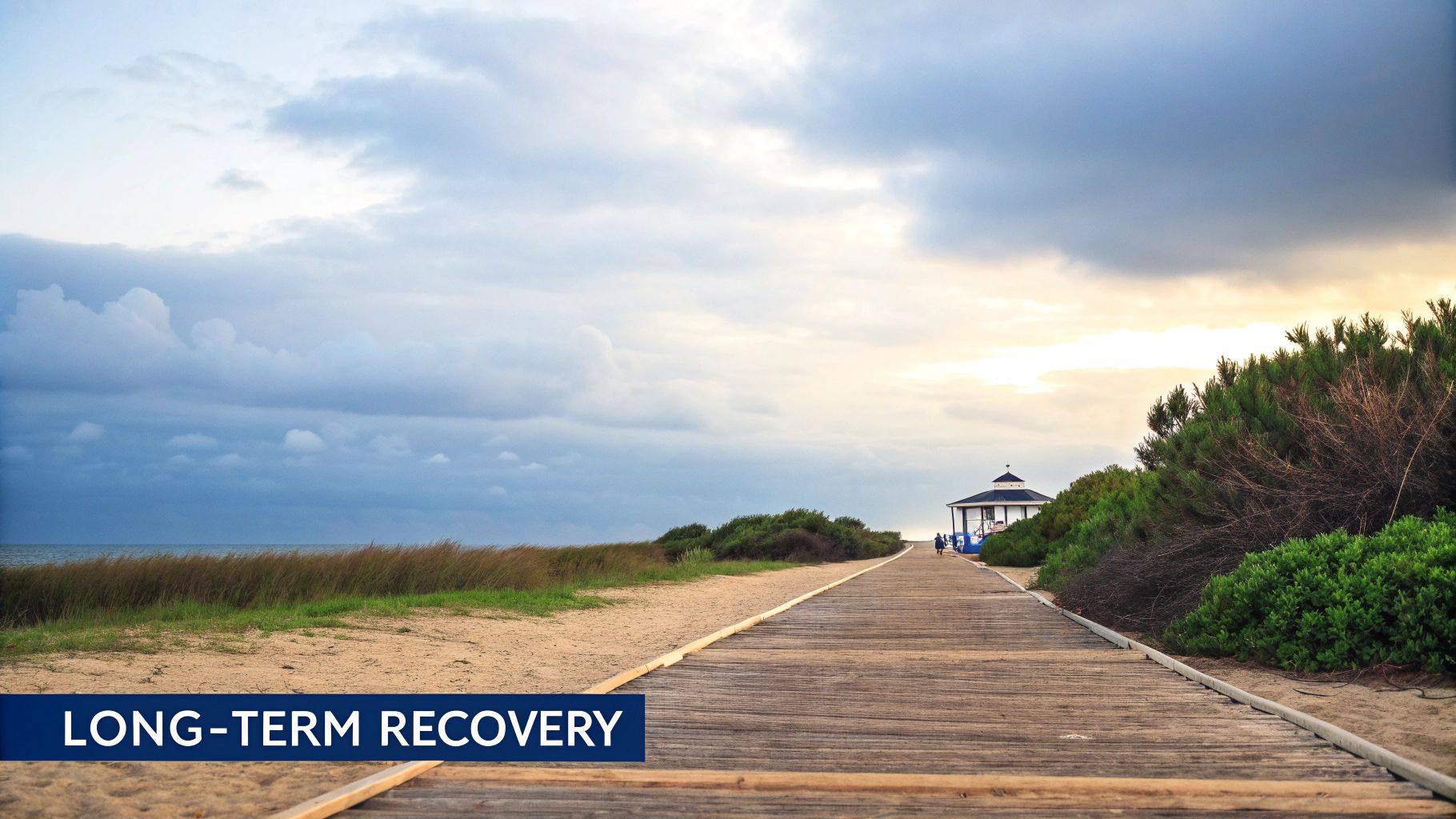 A wooden boardwalk stretches along a sandy beach towards a distant gazebo under a cloudy sky.