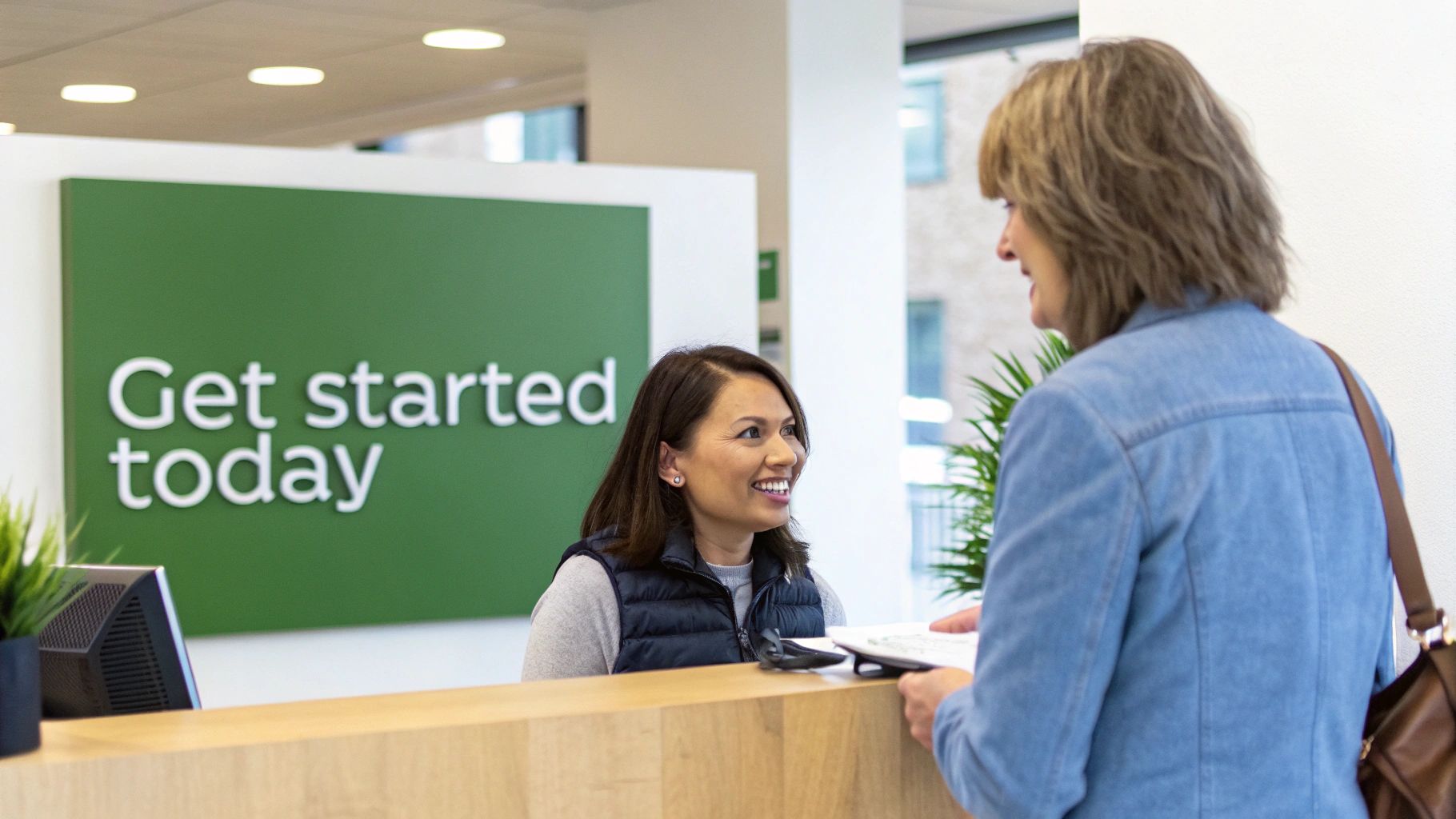 A friendly receptionist smiles at a customer holding documents at a desk with a 'Get started today' sign.