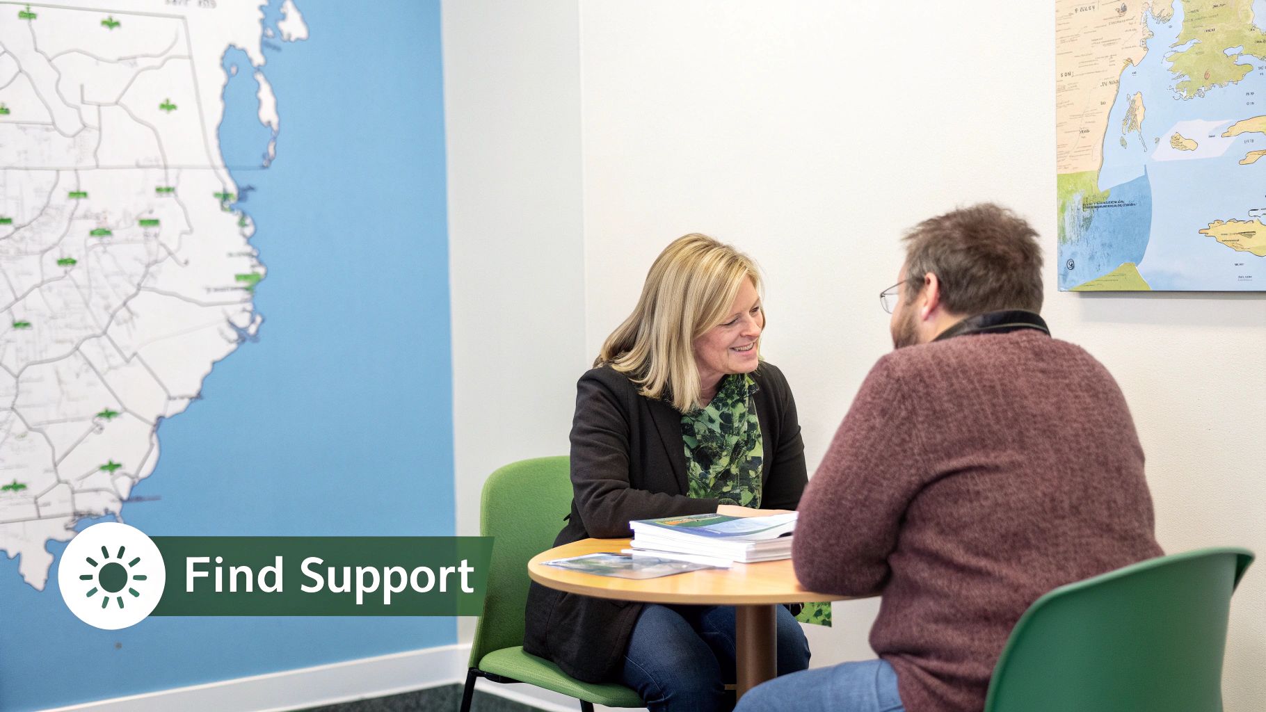 Two people, a man and a woman, sit at a table in a bright room, engaged in a supportive conversation.