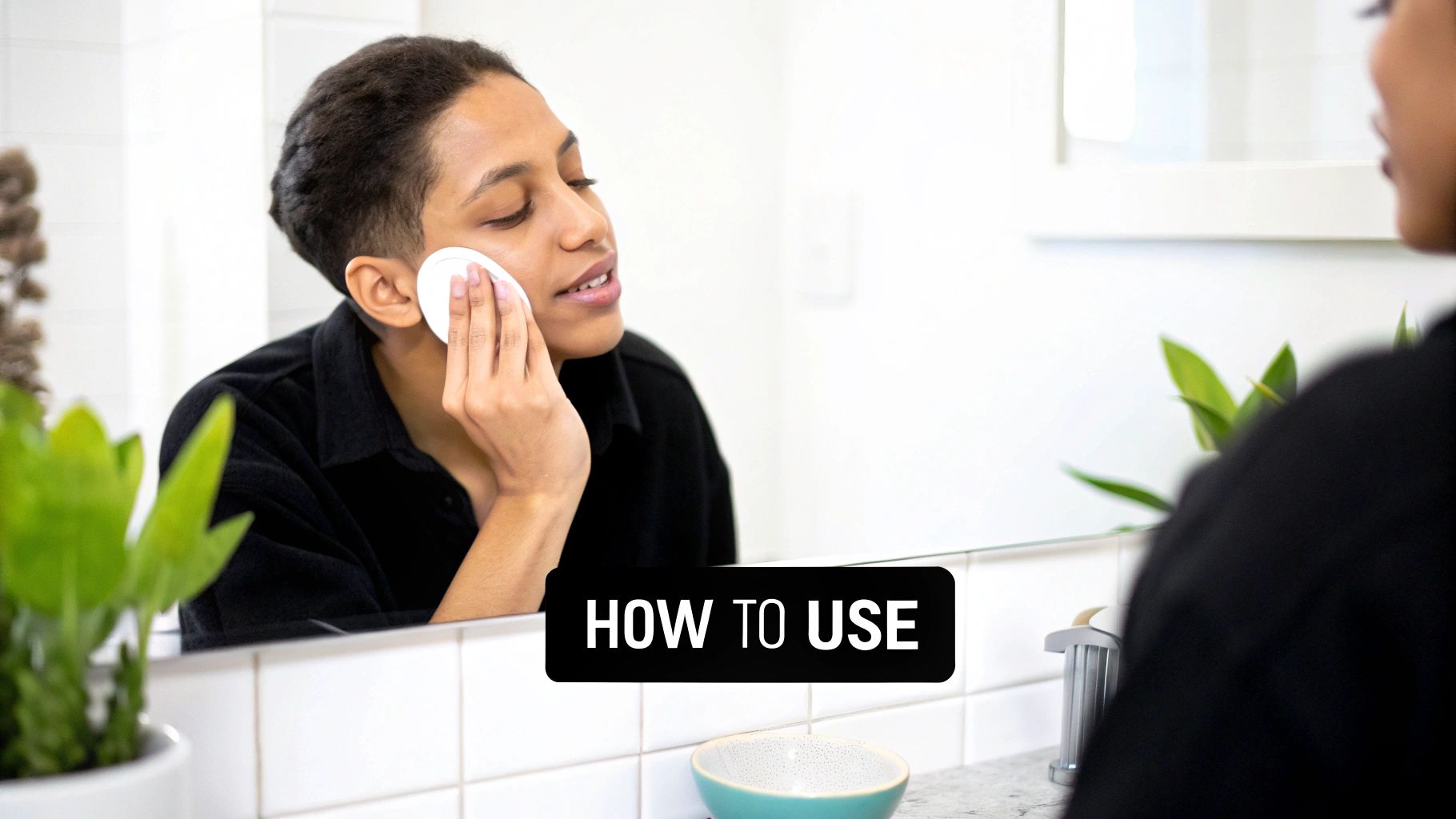 A young adult applies a white salicylic acid pad to their cheek while looking in a bathroom mirror.