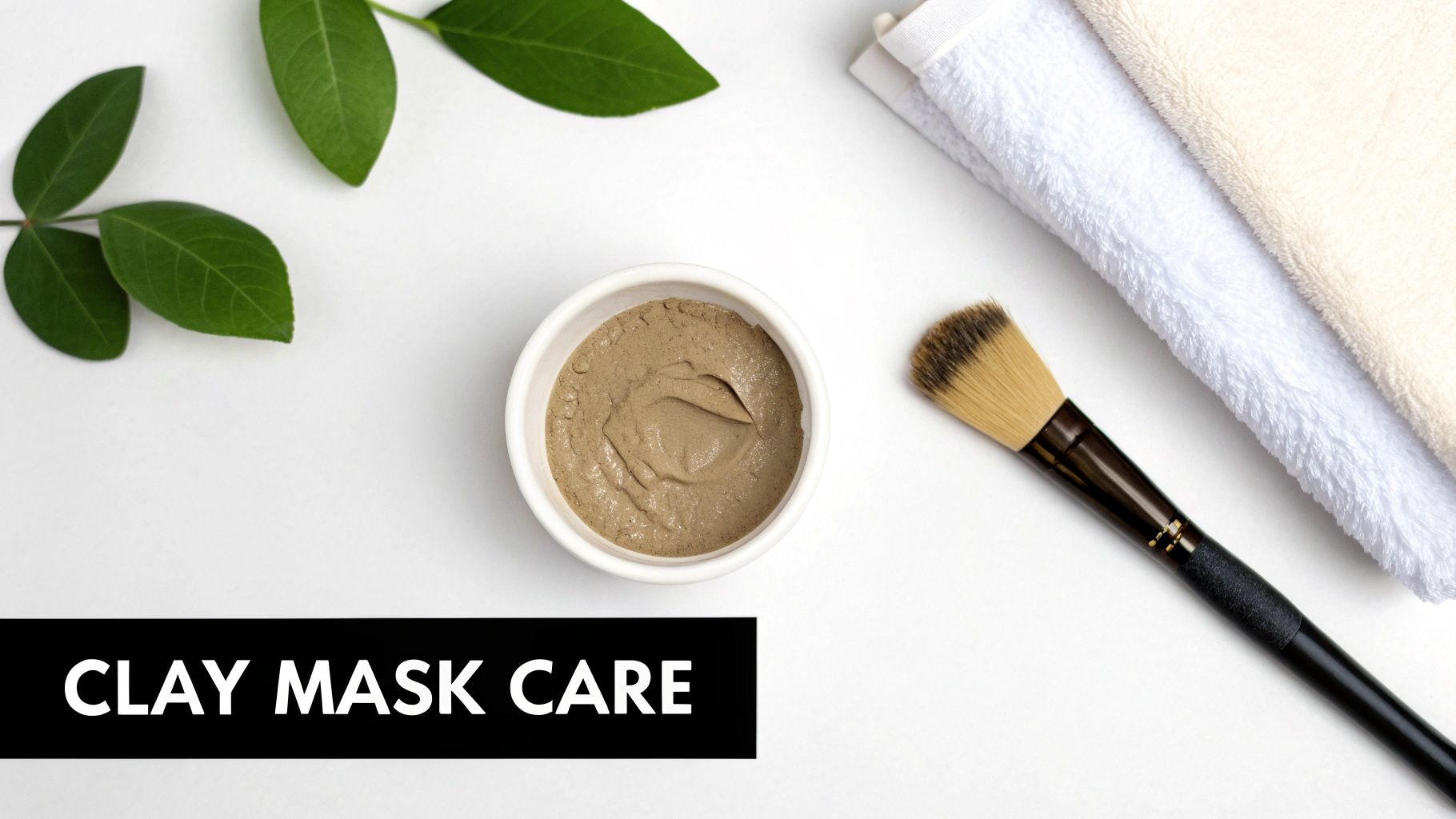 Overhead view of a clay mask in a white bowl, application brush, folded towels, and green leaves.