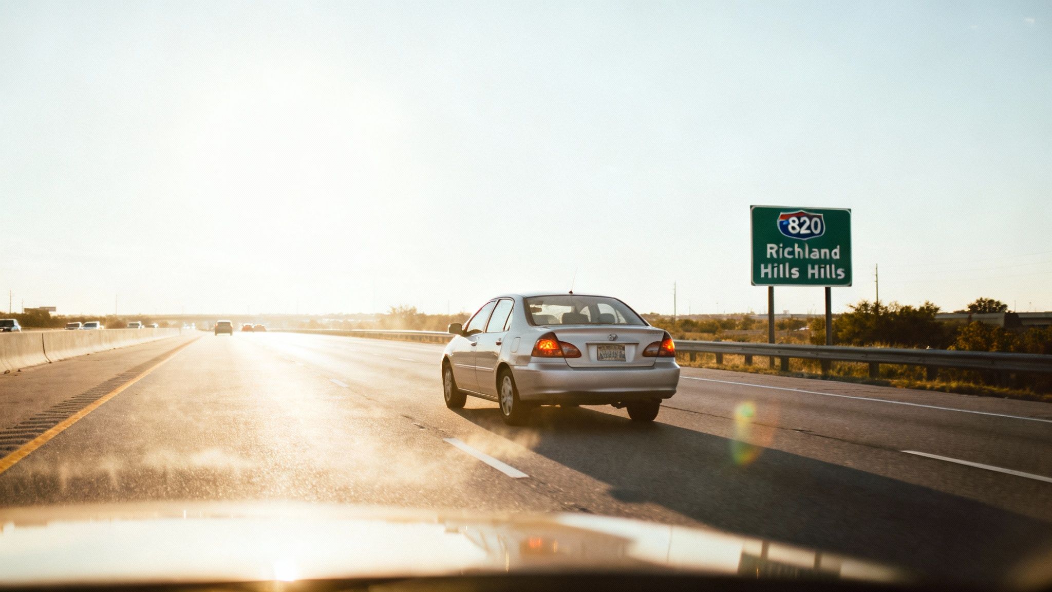 A silver car drives on a multi-lane highway towards a bright sunset, passing a sign for Richland Hills.