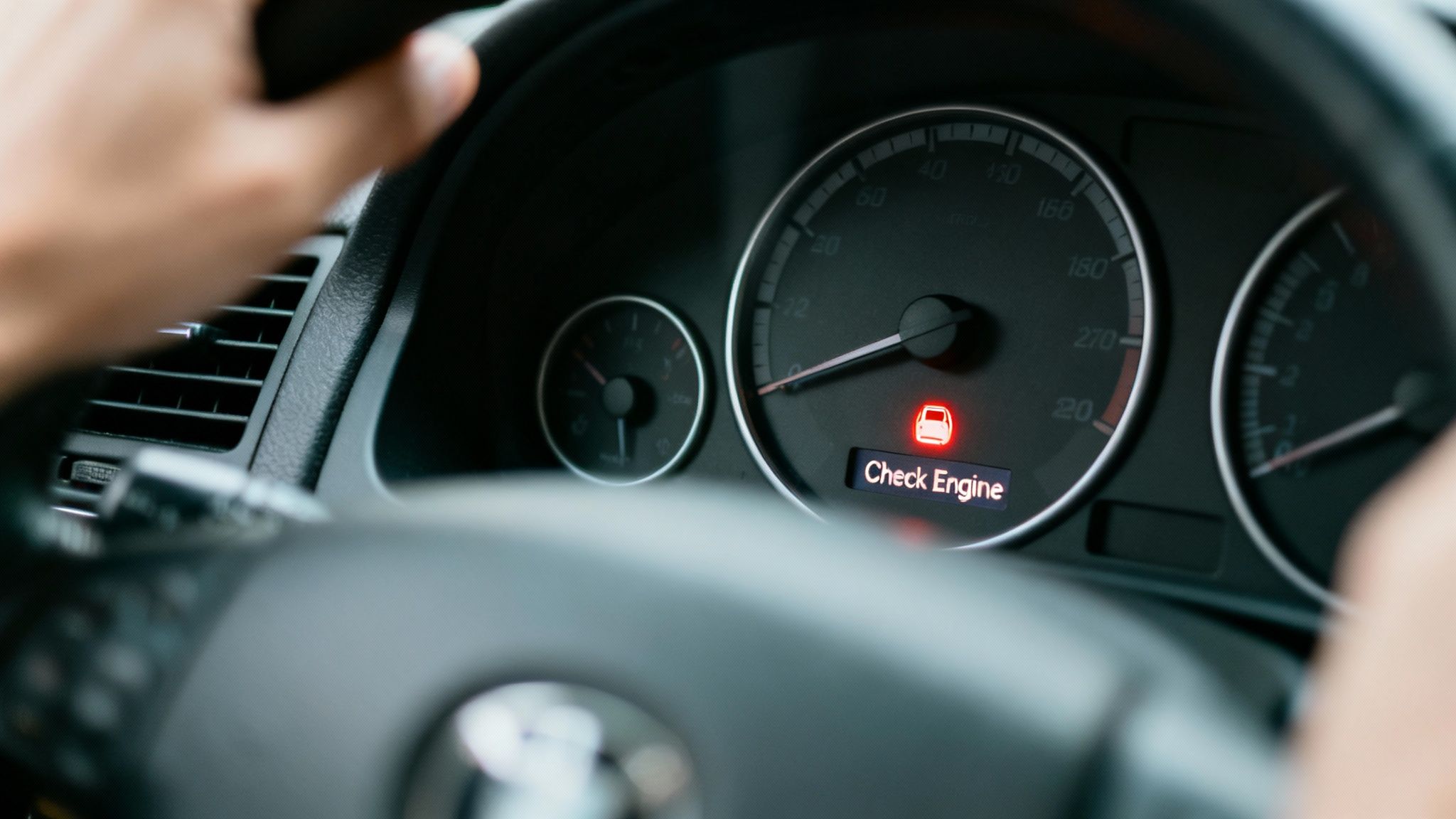 A red 'Check Engine' light is illuminated on a car's dashboard, with a driver's hands on the steering wheel.