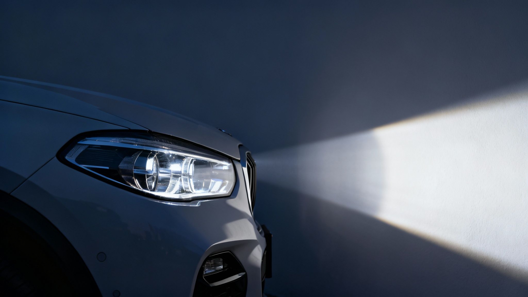 Close-up of a modern white car's illuminated LED headlight casting a bright beam on a dark wall.