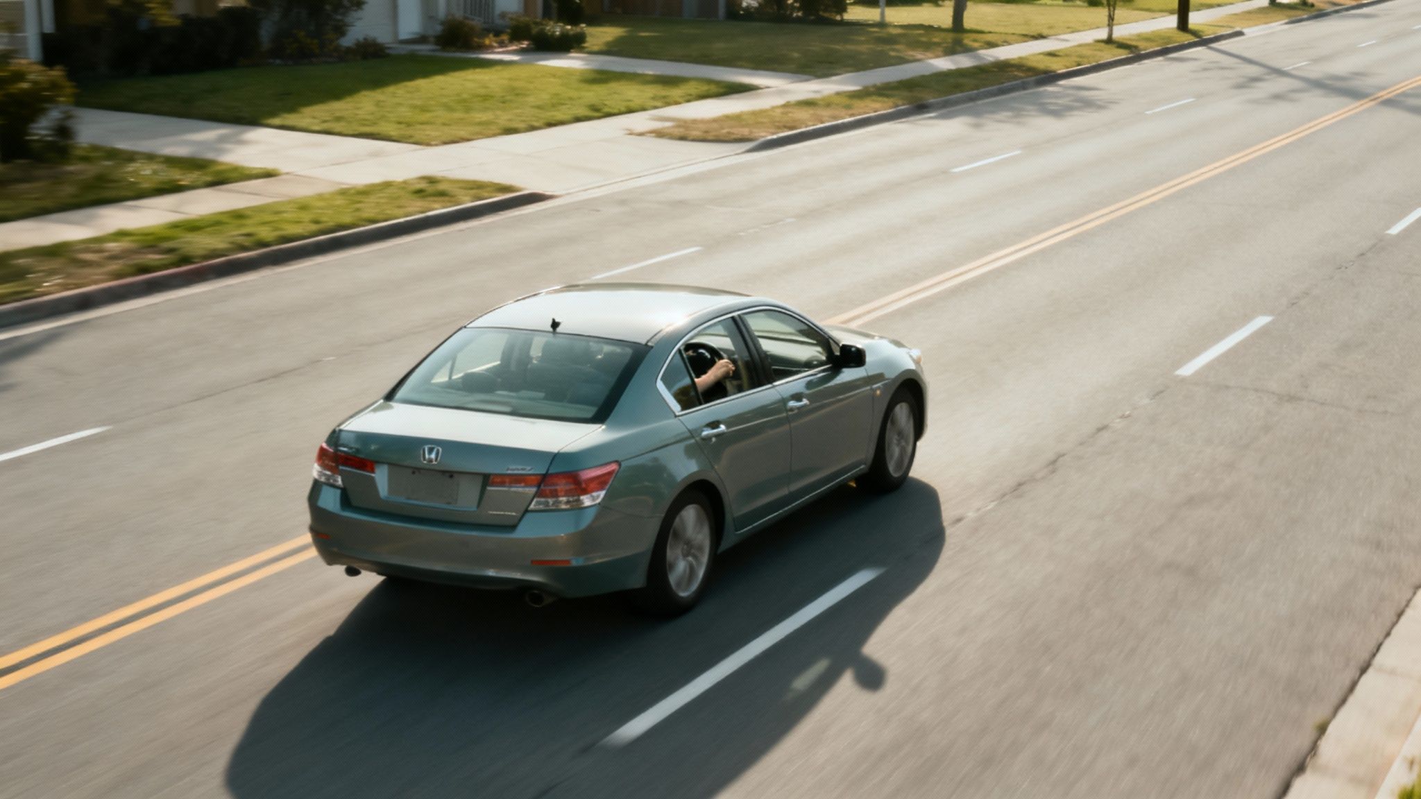 A teal car drives on a sunny road with a person's arm out the driver's window.