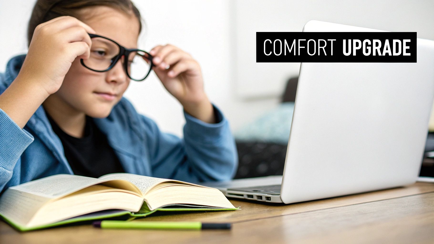 A young girl adjusting her black eyeglasses, studying at a desk with an open book and a laptop.