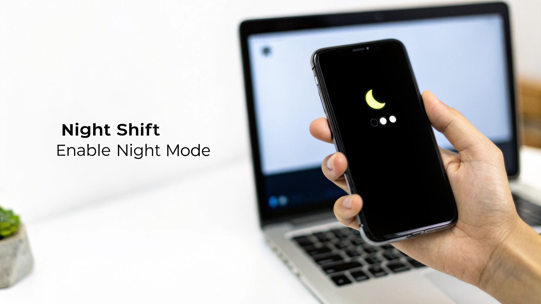 A hand holds a smartphone displaying a crescent moon icon for night mode, with a laptop in the background.
