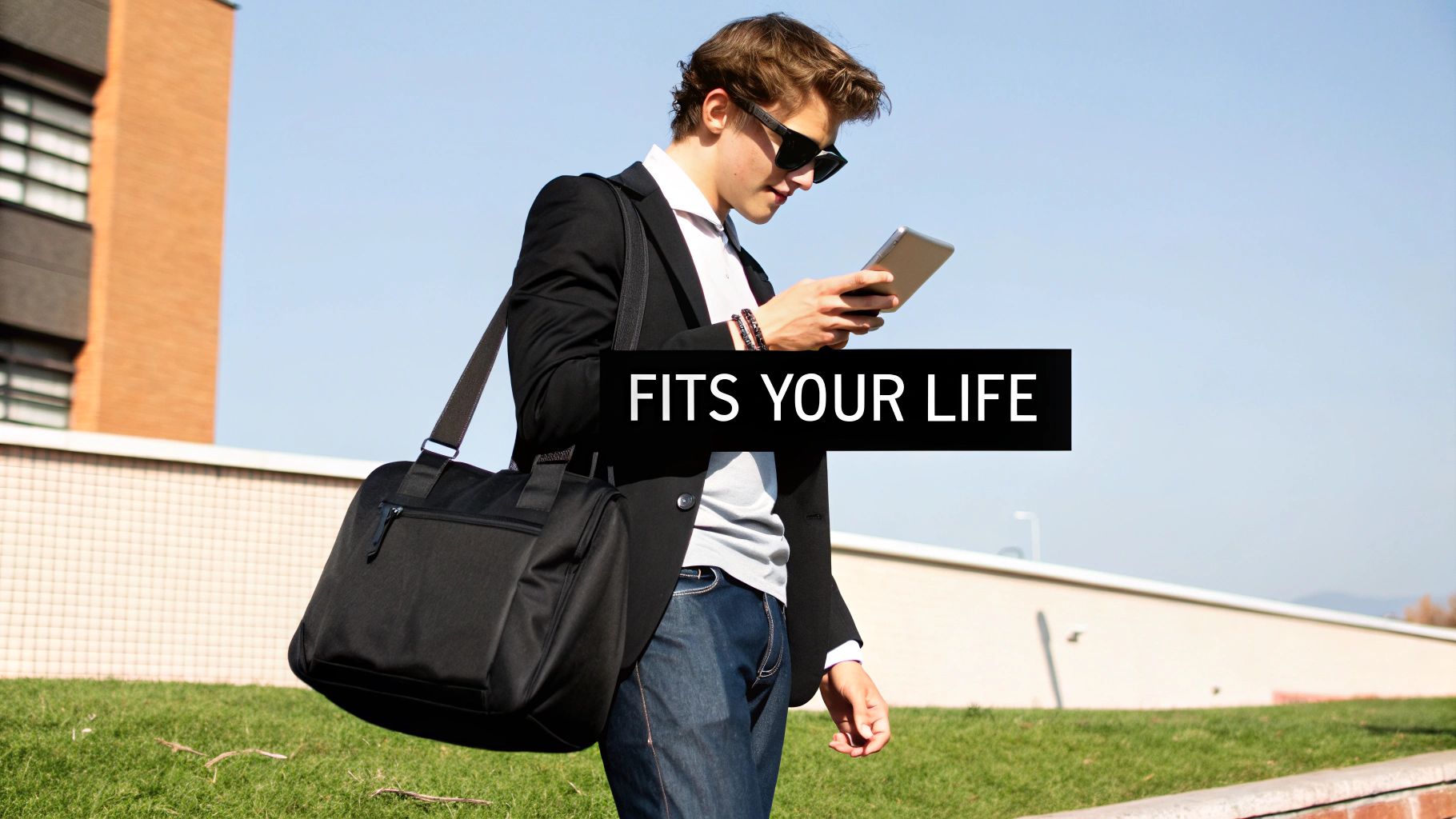 Young man in sunglasses, blazer, and jeans with a messenger bag using a tablet outdoors.