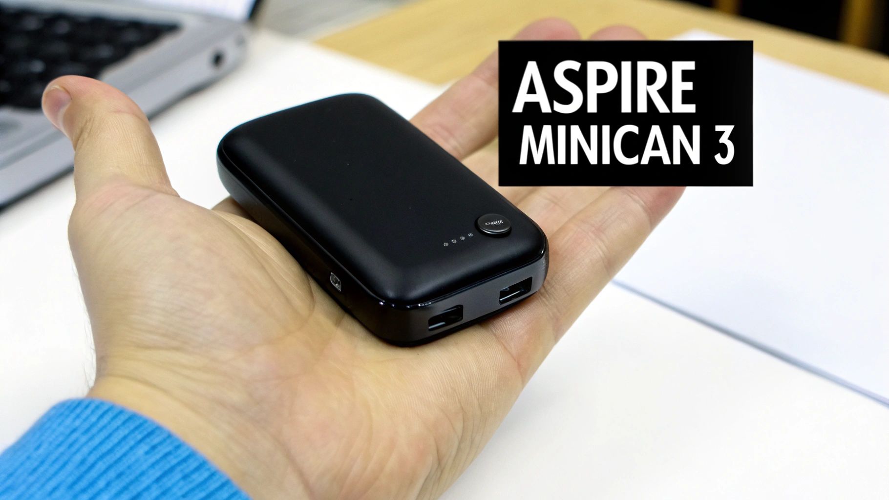 A person holds a black Aspire Minican 3 power bank, with a laptop visible in the background.