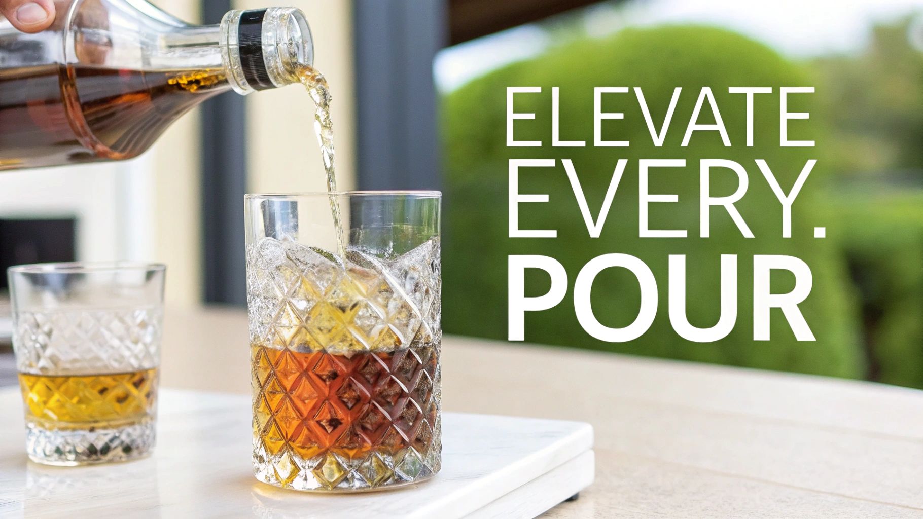 A hand pours amber liquor from a clear bottle into a patterned glass filled with ice.