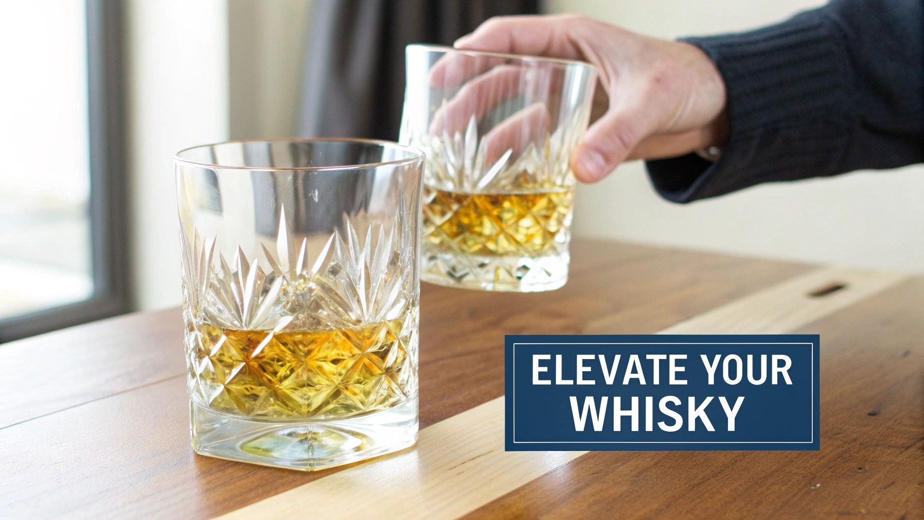 A hand reaches for a crystal glass of whiskey on a wooden table, with another glass nearby.