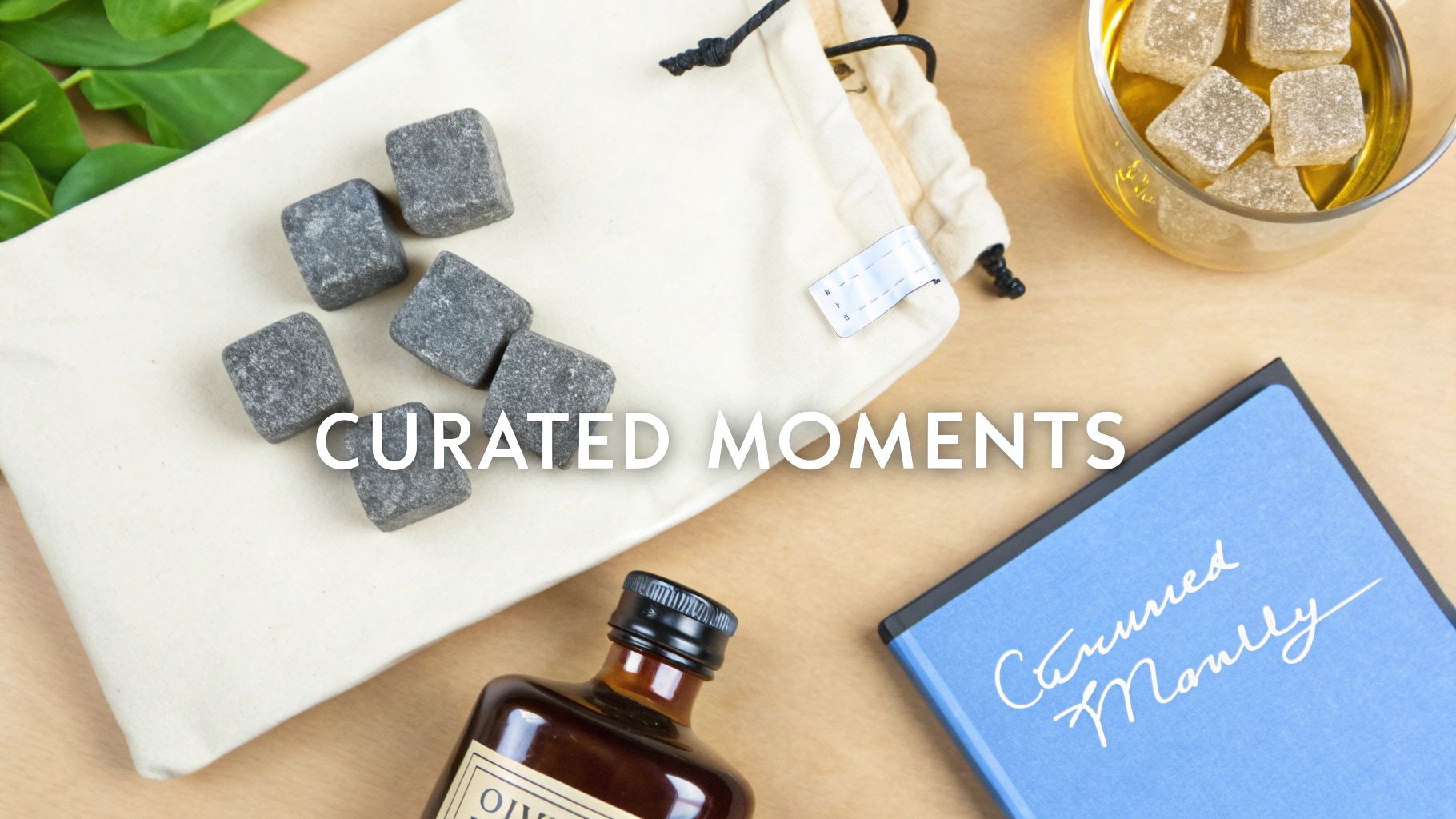 Flat lay of whiskey stones in a canvas pouch, a small bottle, a glass with candied squares, and a blue notebook.