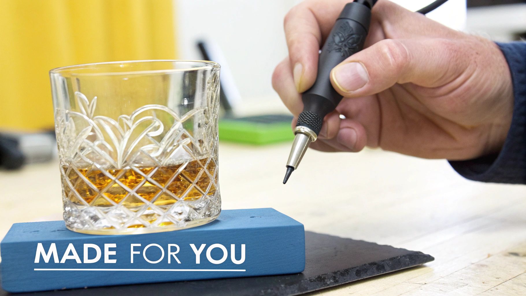 Hand holds an engraving tool over a blue block that reads 'MADE FOR YOU' next to a whiskey glass.