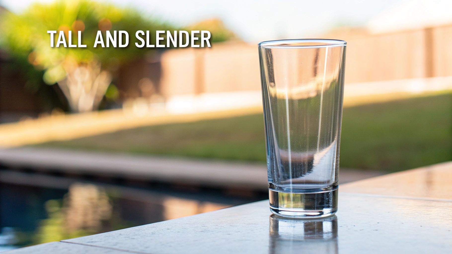 A tall, empty highball glass on a tiled patio next to a swimming pool.